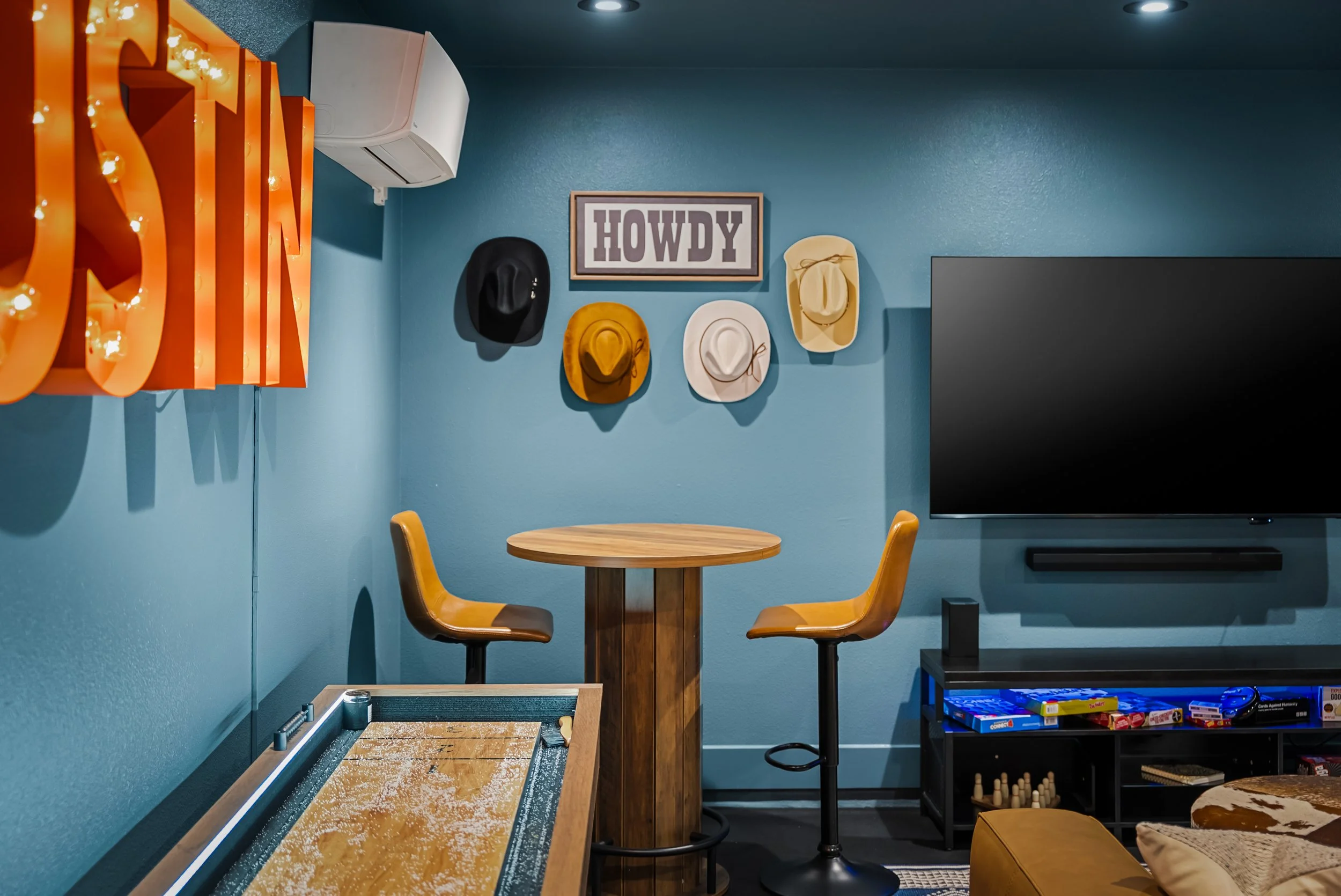 A room with turquoise walls decorated with framed hats and a sign saying 'HOWDY'. There is a wooden table with two brown chairs, a small black shelf with board games, a large TV, and a large illuminated orange sign that reads 'FUN'.