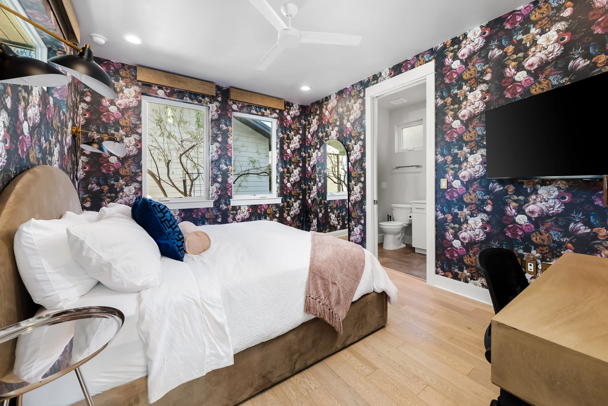 Bedroom with floral patterned wallpaper, a bed with white bedding and blue and beige pillows, windows with trees outside, a television on the wall, a wooden desk, and a small bathroom visible through an open door.