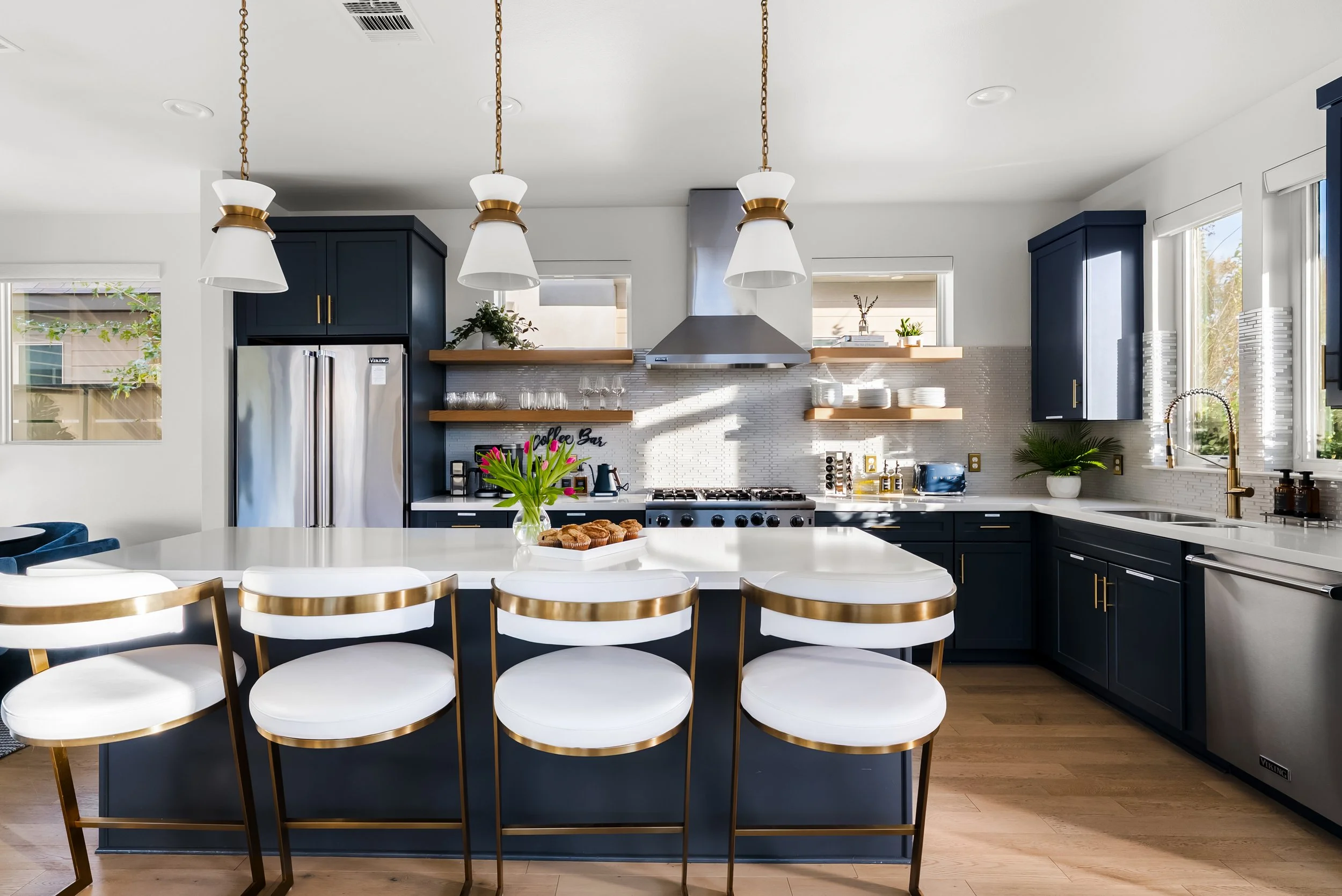Modern kitchen with navy blue cabinets, white countertops, stainless steel appliances, and a kitchen island with white and gold chairs. Pendant lights hang above the island, and there are open shelves with dishes and glassware. Natural light streams 