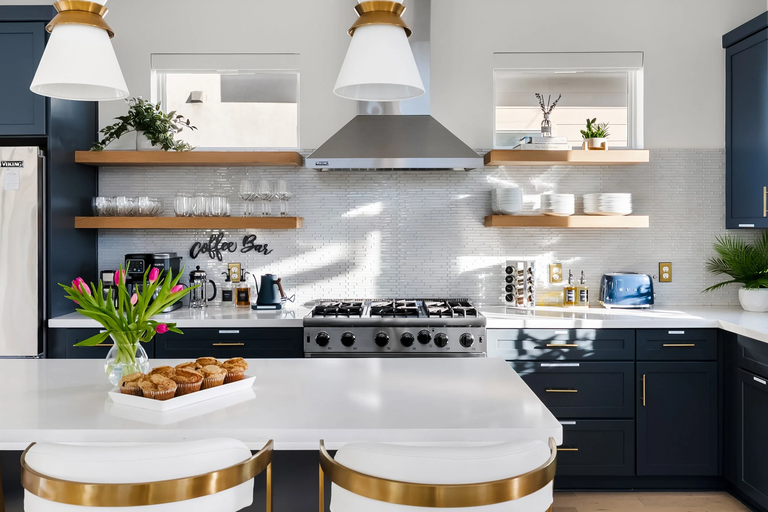 Modern kitchen with navy blue cabinets, white countertops, open wooden shelves, black appliances, a stove, a range hood, and decorative plants, with sunlight streaming in.