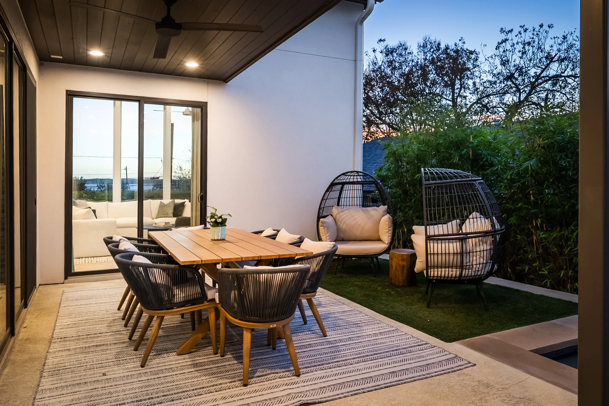 Outdoor patio area with dining table and chairs, two hanging egg chairs with cushions, a wooden side table, greenery bushes, and a view of the evening sky through the trees.