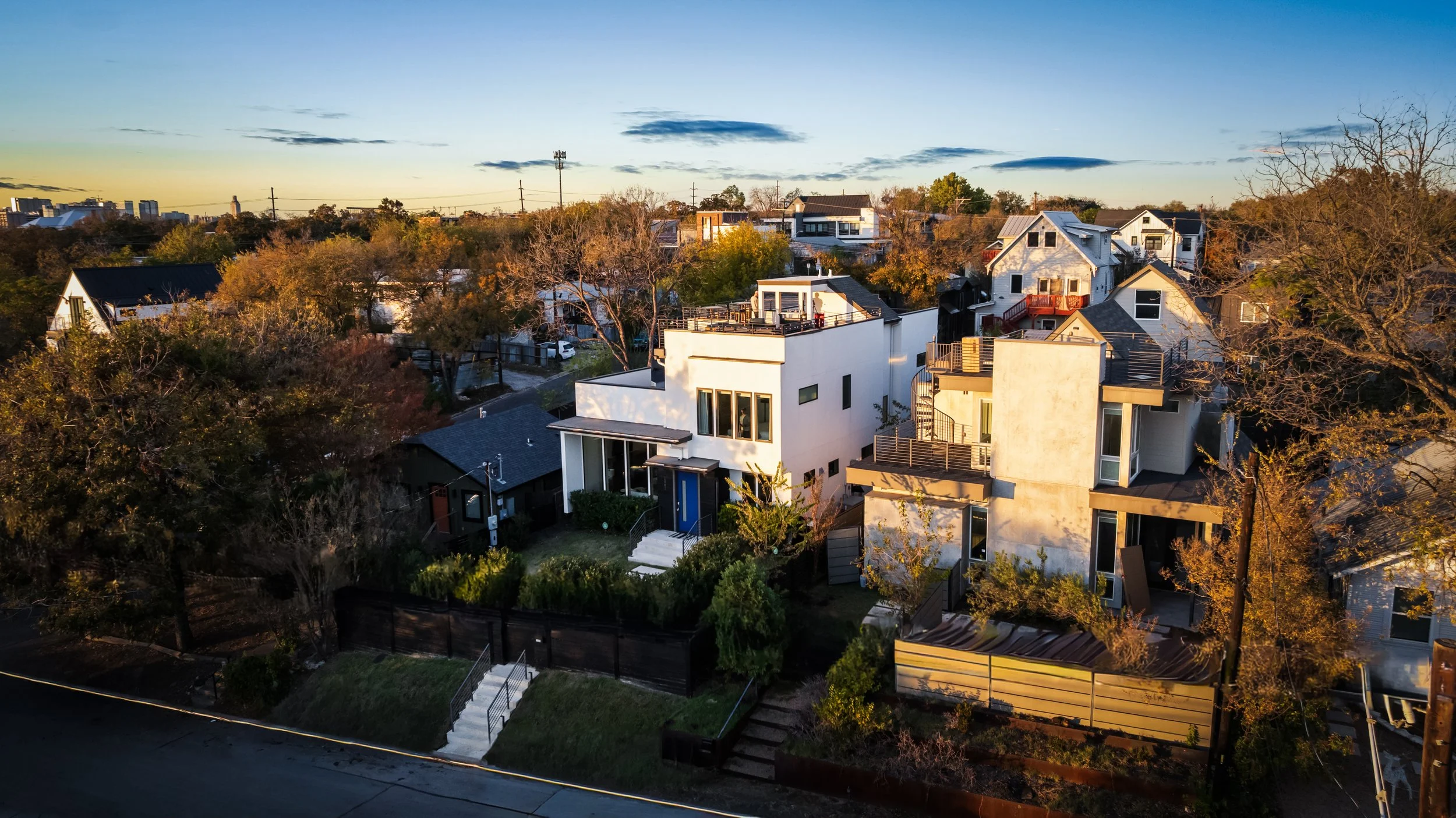 Aerial view of modern residential neighborhood at sunset, featuring contemporary houses with balconies and terraces, surrounded by trees and a fenced yard.
