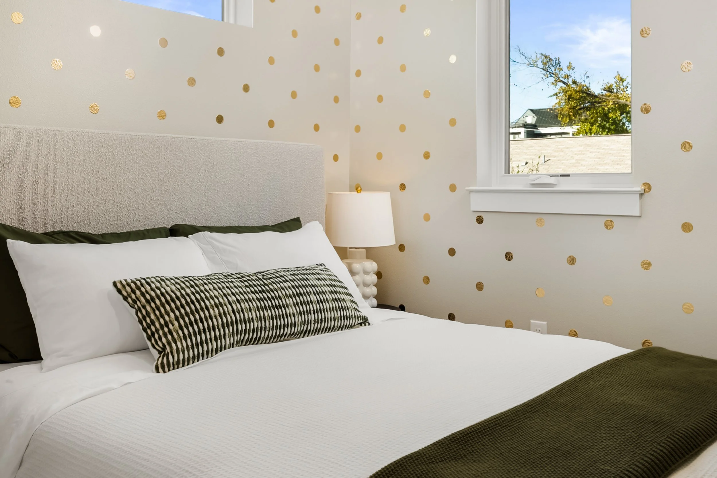 A cozy bedroom with a bed having white bedding, a black and white striped pillow, and dark green pillows. There is a white lamp with a textured base on a nightstand beside the bed. The wall behind the bed is decorated with gold polka dots, and a window shows a clear blue sky and trees outside.