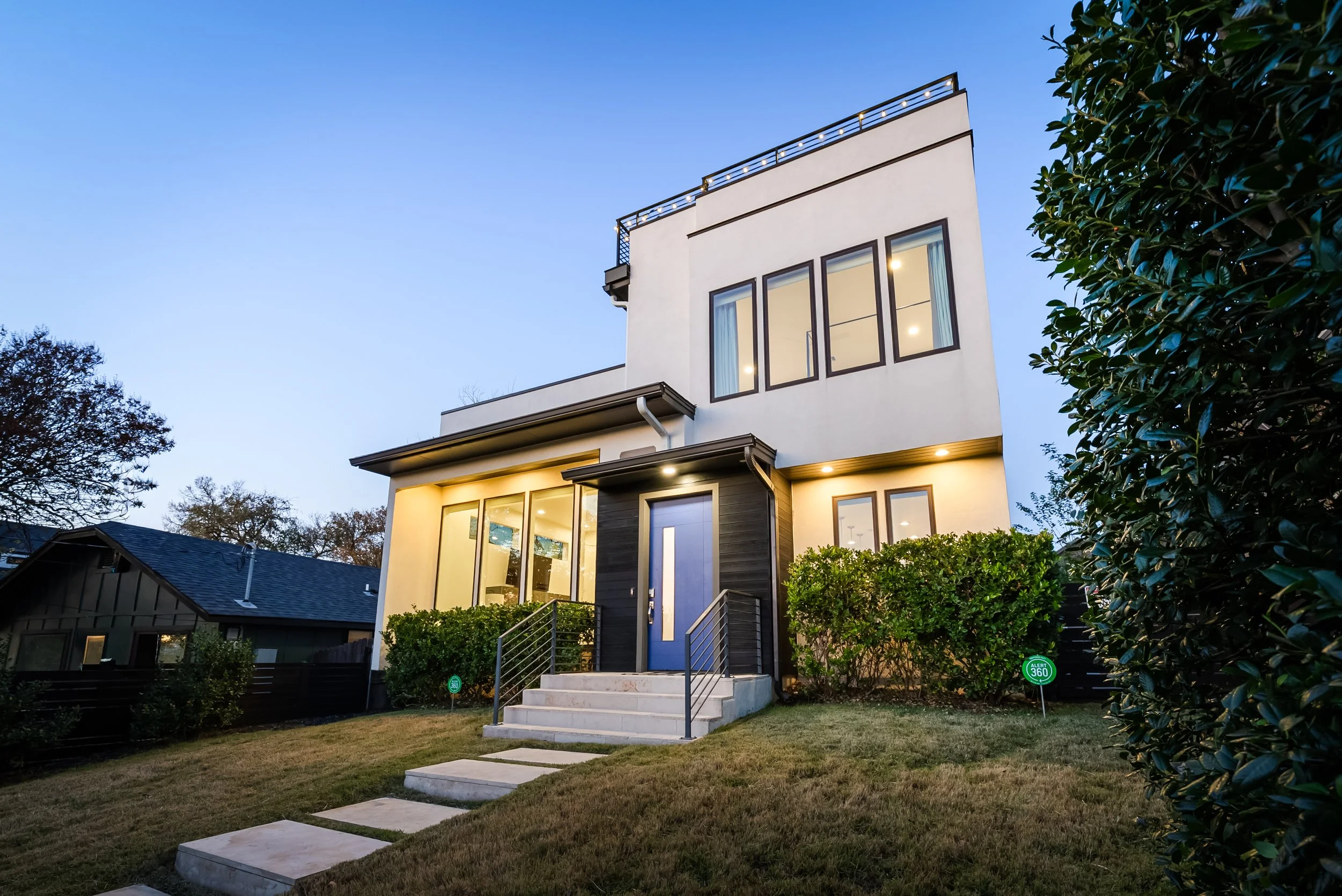 Modern multi-story house with large windows, illuminated from inside, surrounded by a well-maintained lawn, bushes, and trees, with a pathway leading to the front door, during dusk.