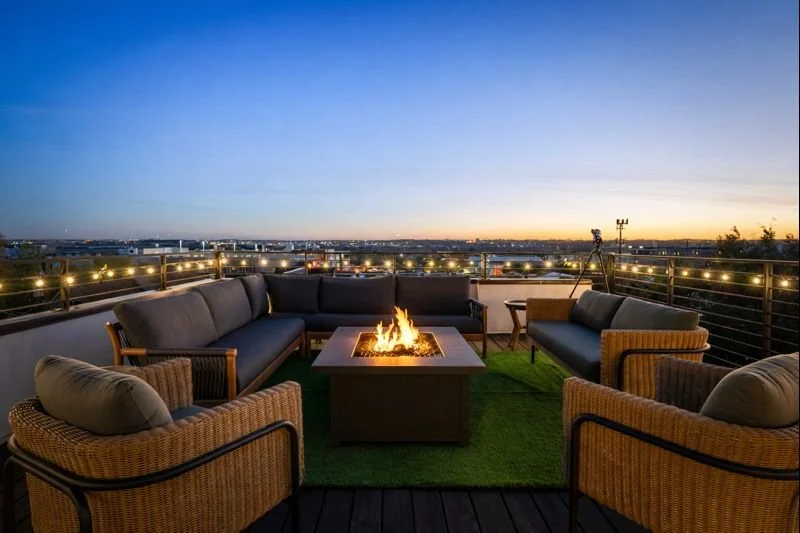 Rooftop patio with outdoor sofas and chairs, string lights, a green rug, a telescope, and a view of the city skyline at sunset.