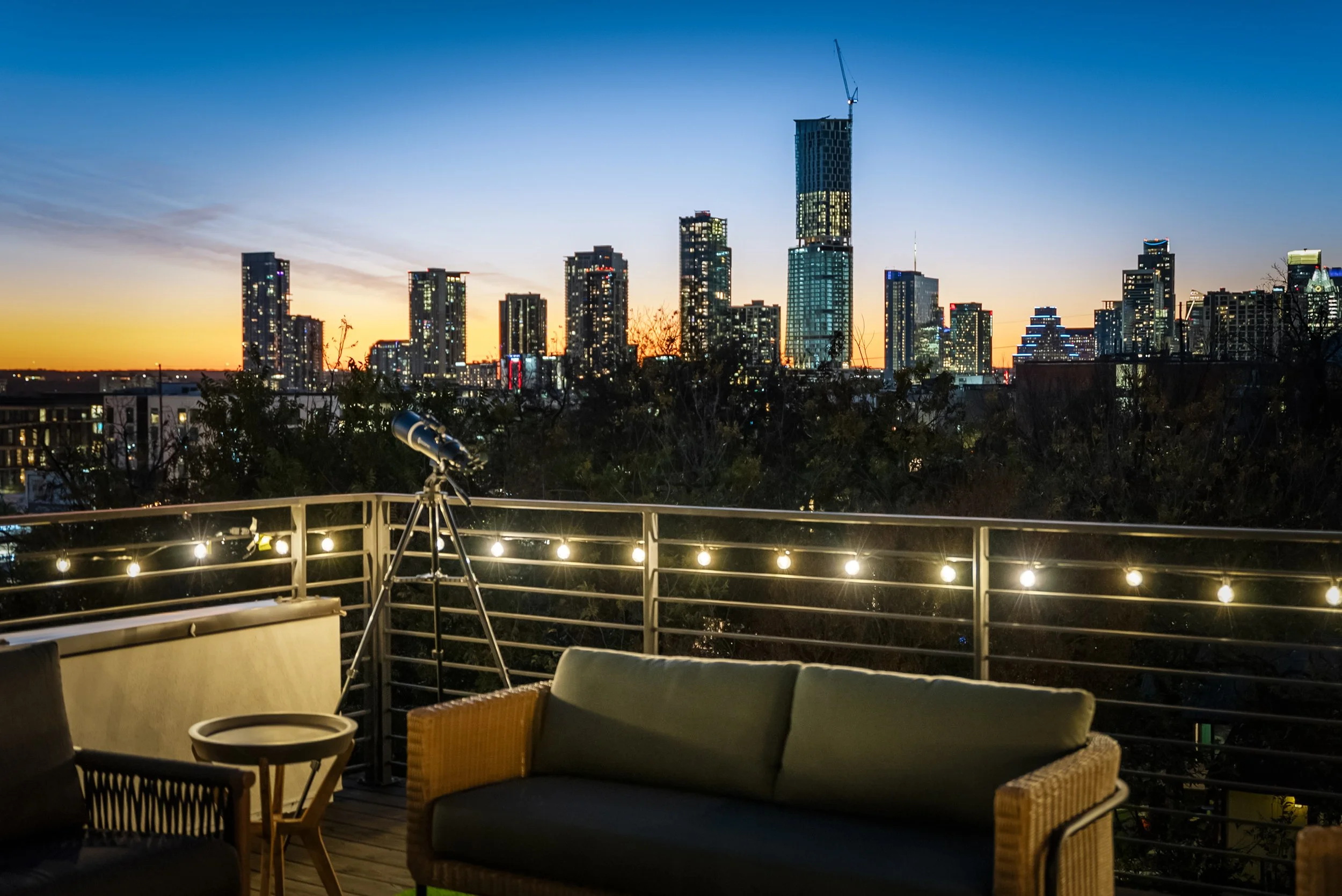 City skyline at dusk seen from a rooftop patio, with outdoor furniture, string lights, and a telescope.