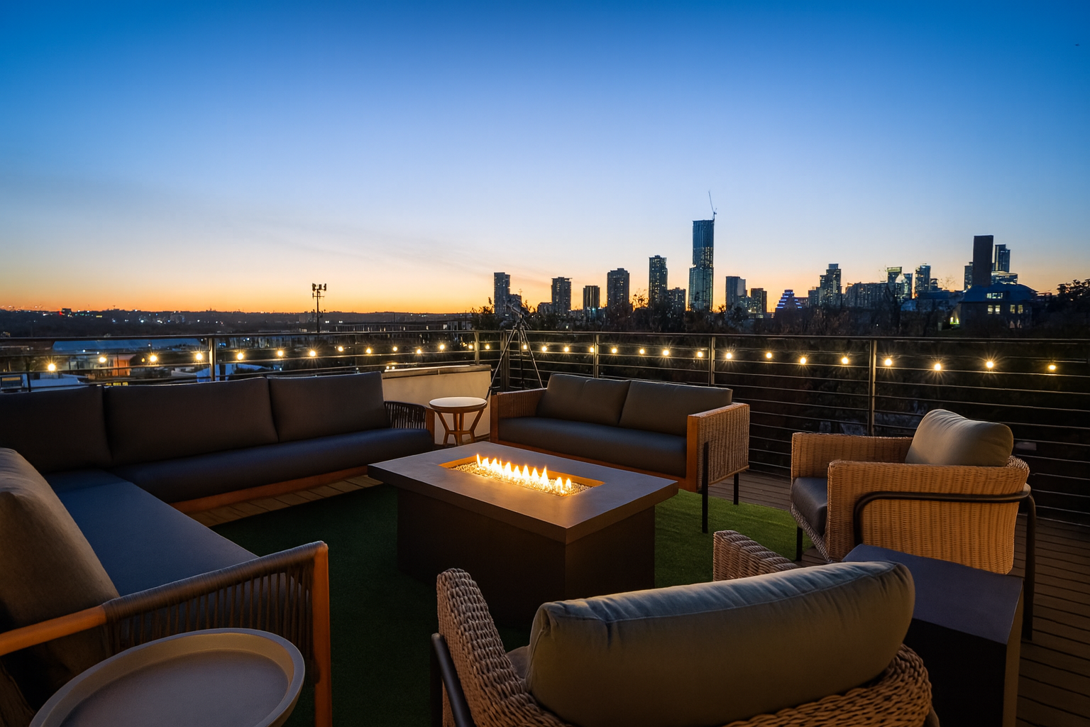 Rooftop lounge with outdoor sofas around a fire pit, string lights, city skyline at sunset in the background.