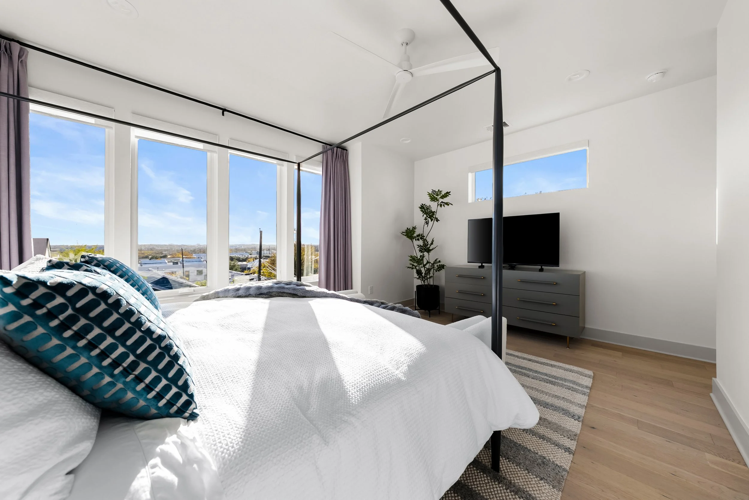 Modern bedroom with a white bed, blue patterned pillows, large windows with a view of the outdoors, a black canopy bed frame, a gray dresser with a TV, a potted plant, and a wooden floor.