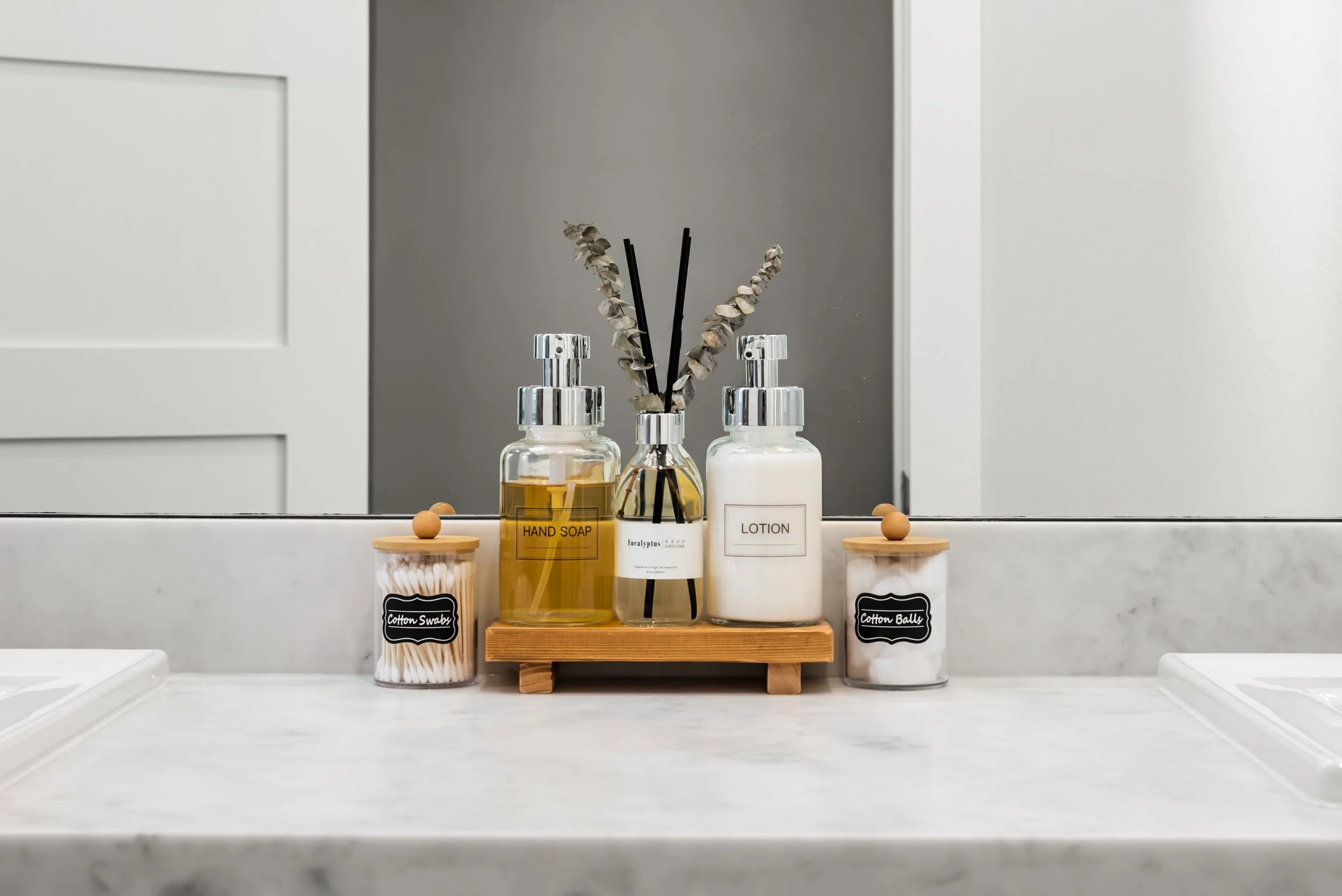 Bathroom countertop with two soap dispensers labeled Hand Soap and Lotion, two cotton swabs jars, a reed diffuser, and a mirror reflecting part of the room.
