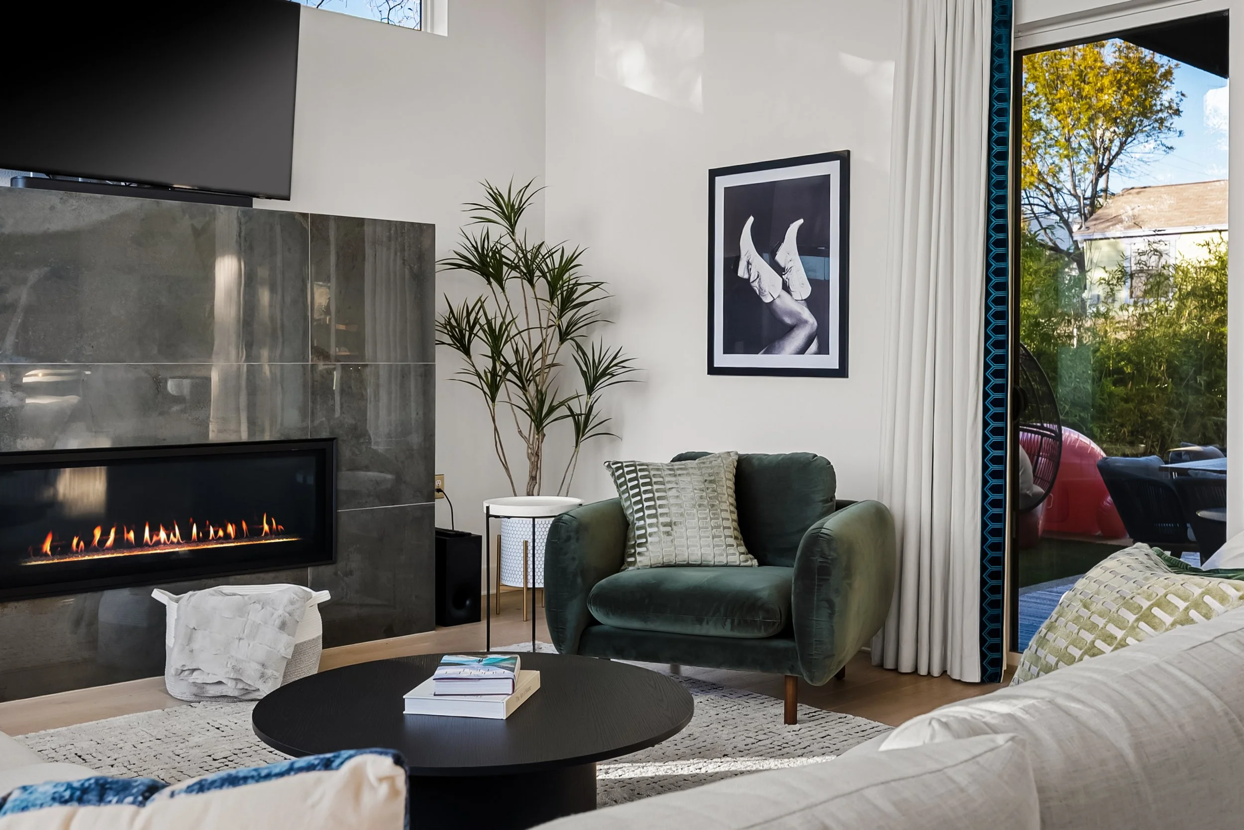 Living room with a fireplace, green armchair, potted plant, framed black-and-white art, round coffee table with books, and sliding glass door revealing outdoor patio and trees.