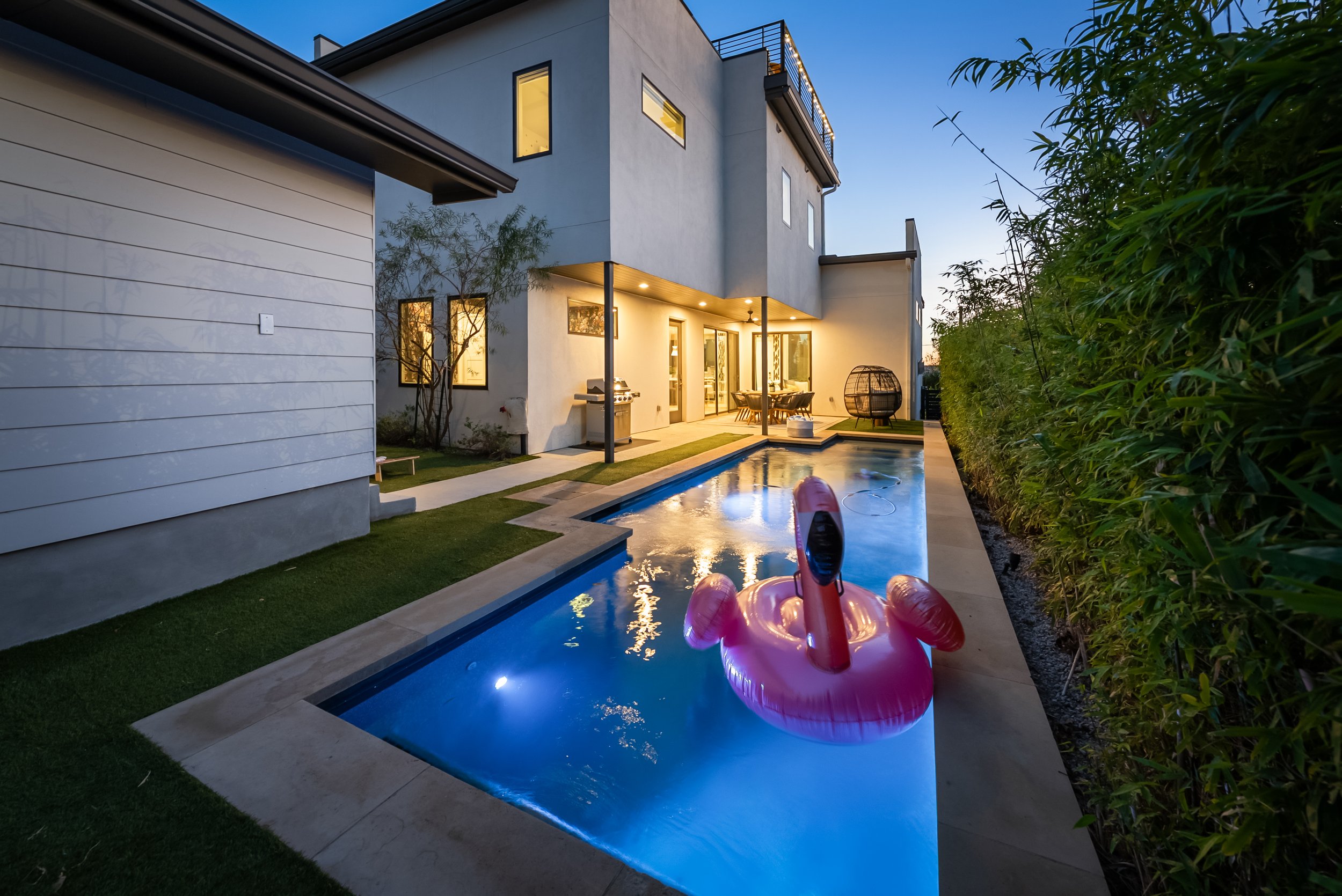 Modern house backyard with lit swimming pool, pink inflatable flamingo float, outdoor dining table, grill, and surrounded by green hedges at dusk.