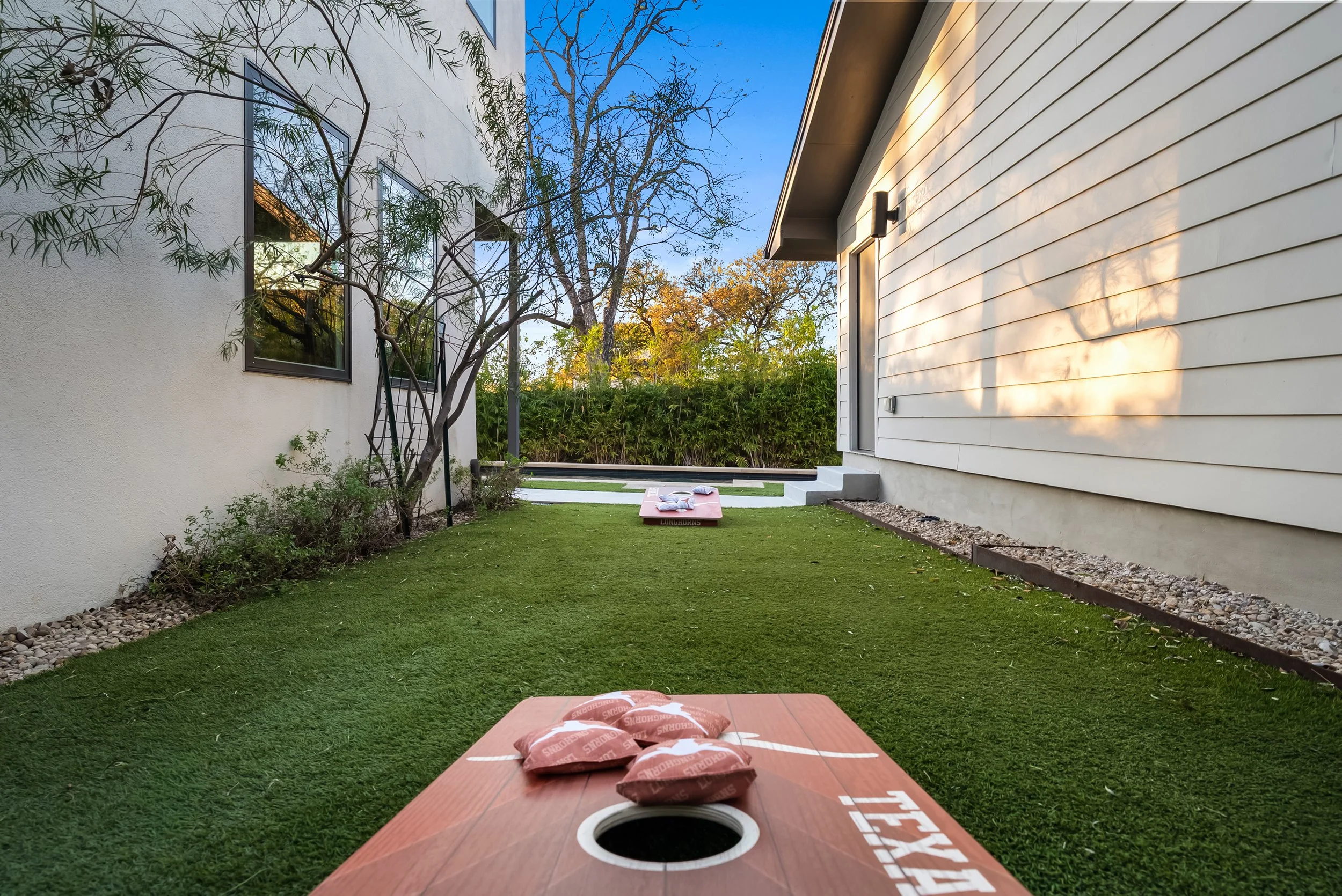 Backyard cornhole game setup on artificial grass with two boards, one in the foreground and one in the background. Residential houses with siding and large windows are on either side, and trees with autumn leaves are in the background under a clear blue sky.