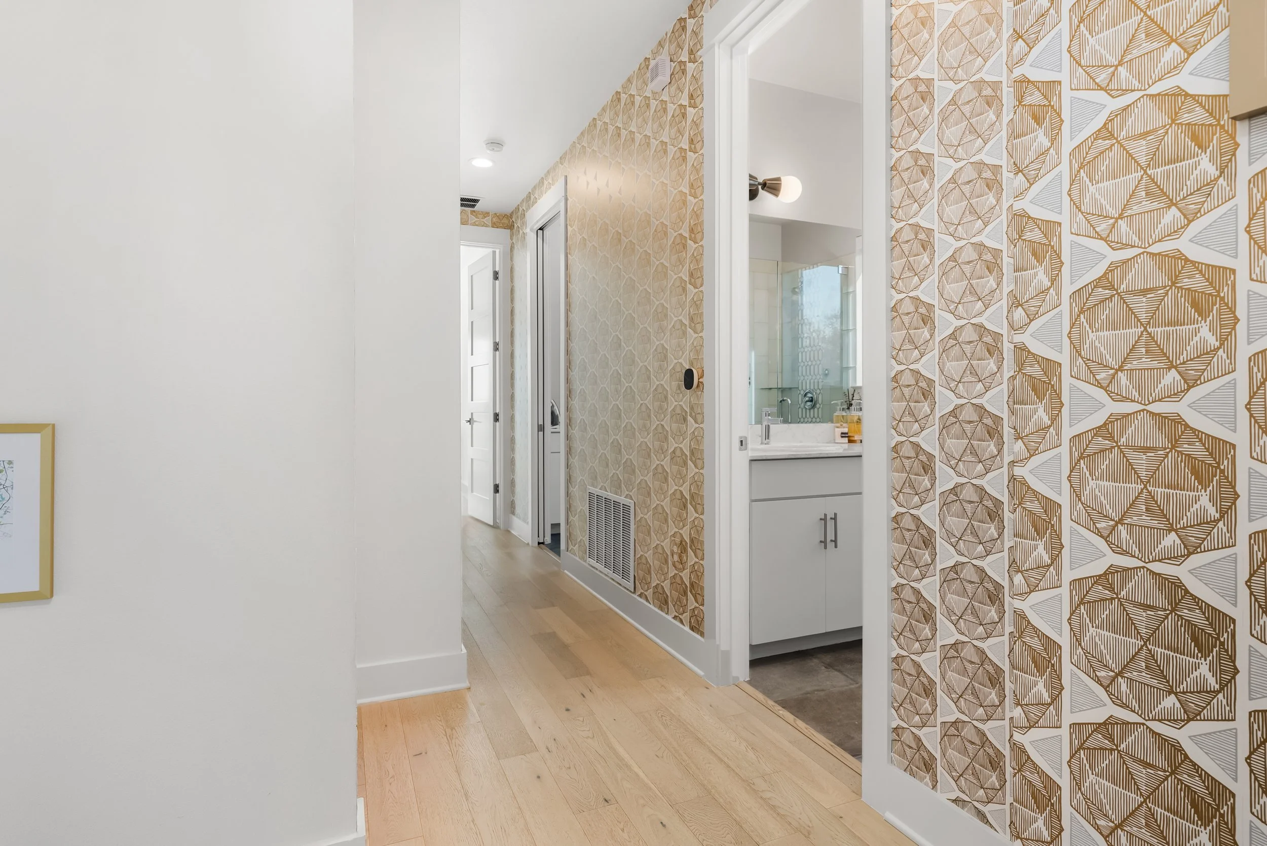 A hallway with light wood floors, white walls, and a bathroom on the right with patterned wallpaper featuring geometric designs in gold and white. Further down the hall are two white doors and a closed white vent.