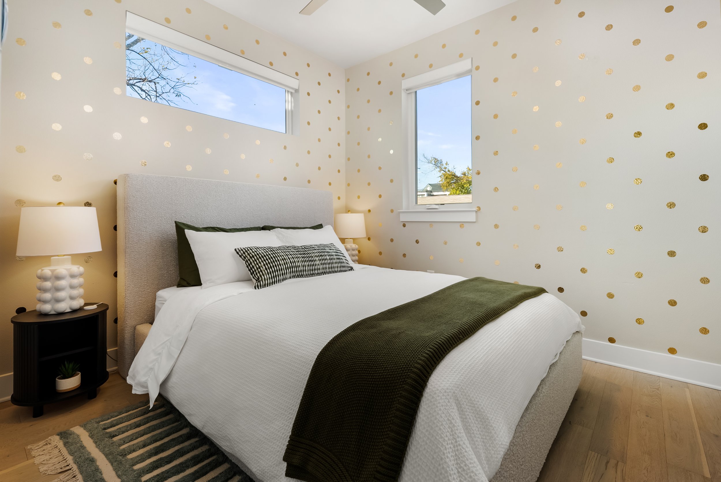A bedroom with a double bed with white bedding, green throw, and pillows, against a wall with white wallpaper decorated with gold polka dots, and two windows showing a blue sky and trees outside.