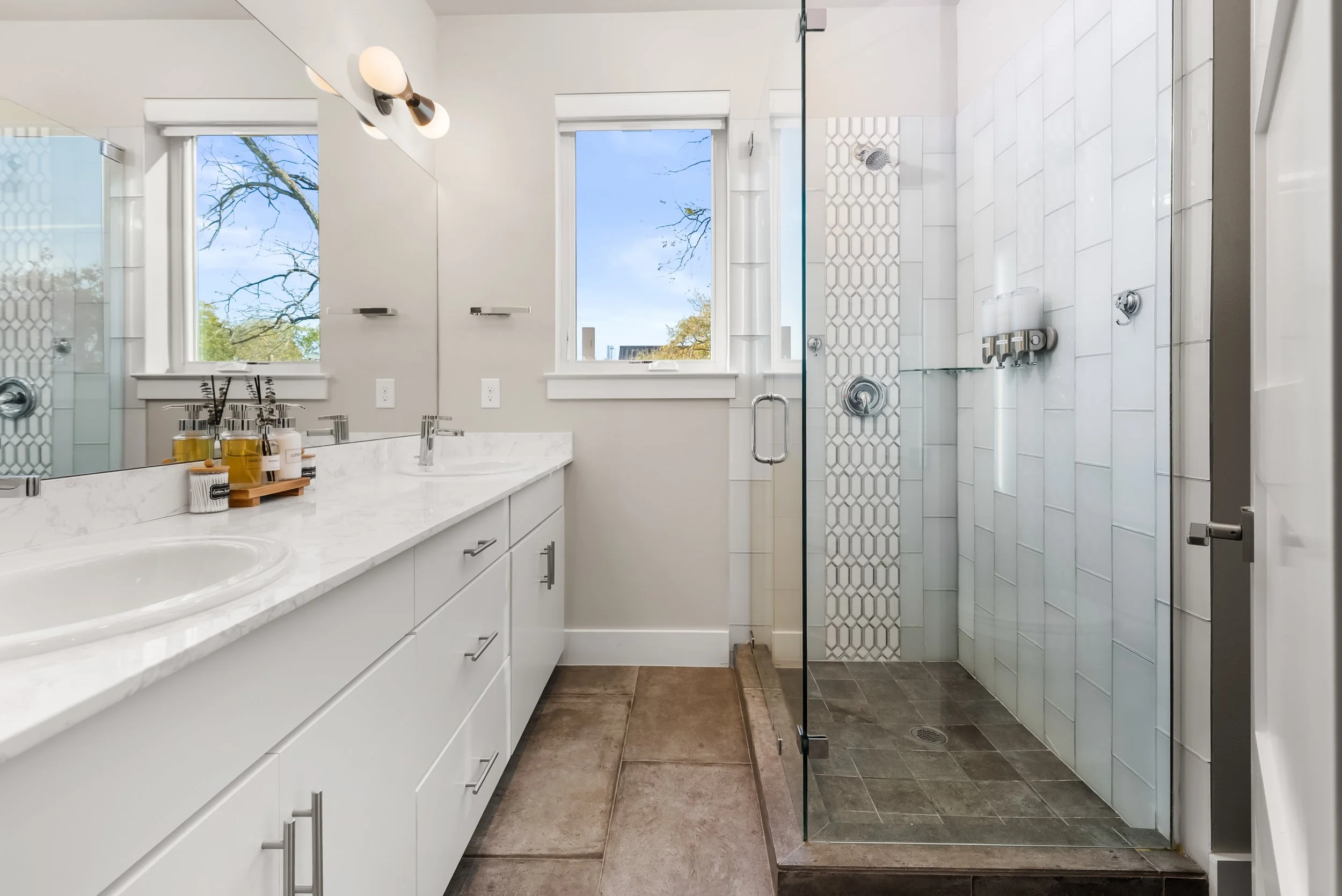 Modern bathroom featuring a white vanity with marble countertop, two windows showing trees outside, and a walk-in shower with glass door and white tiles.