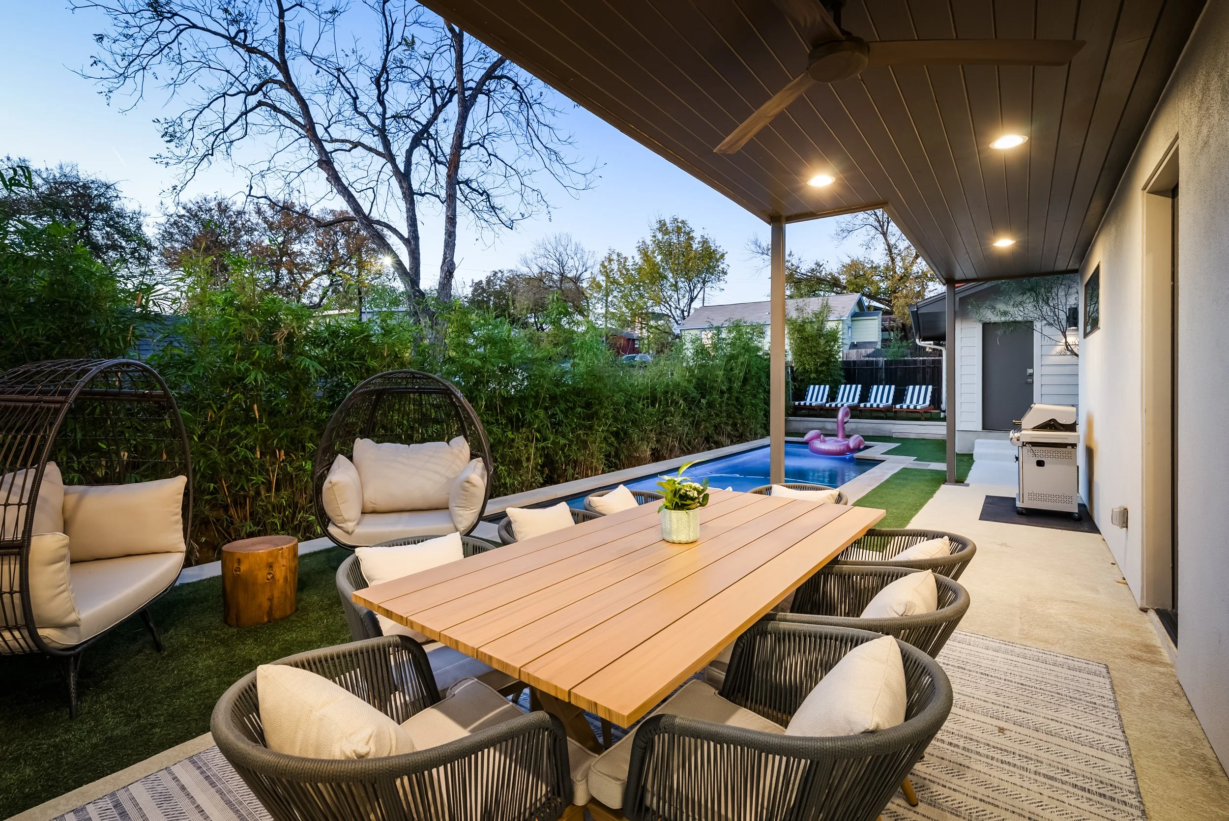 Outdoor patio with a wooden dining table, multiple cushioned chairs, woven hanging chairs, a small side table, and a grill, next to a pool with an inflatable swan, surrounded by greenery and trees, under a covered porch with ceiling lights and a ceiling fan.