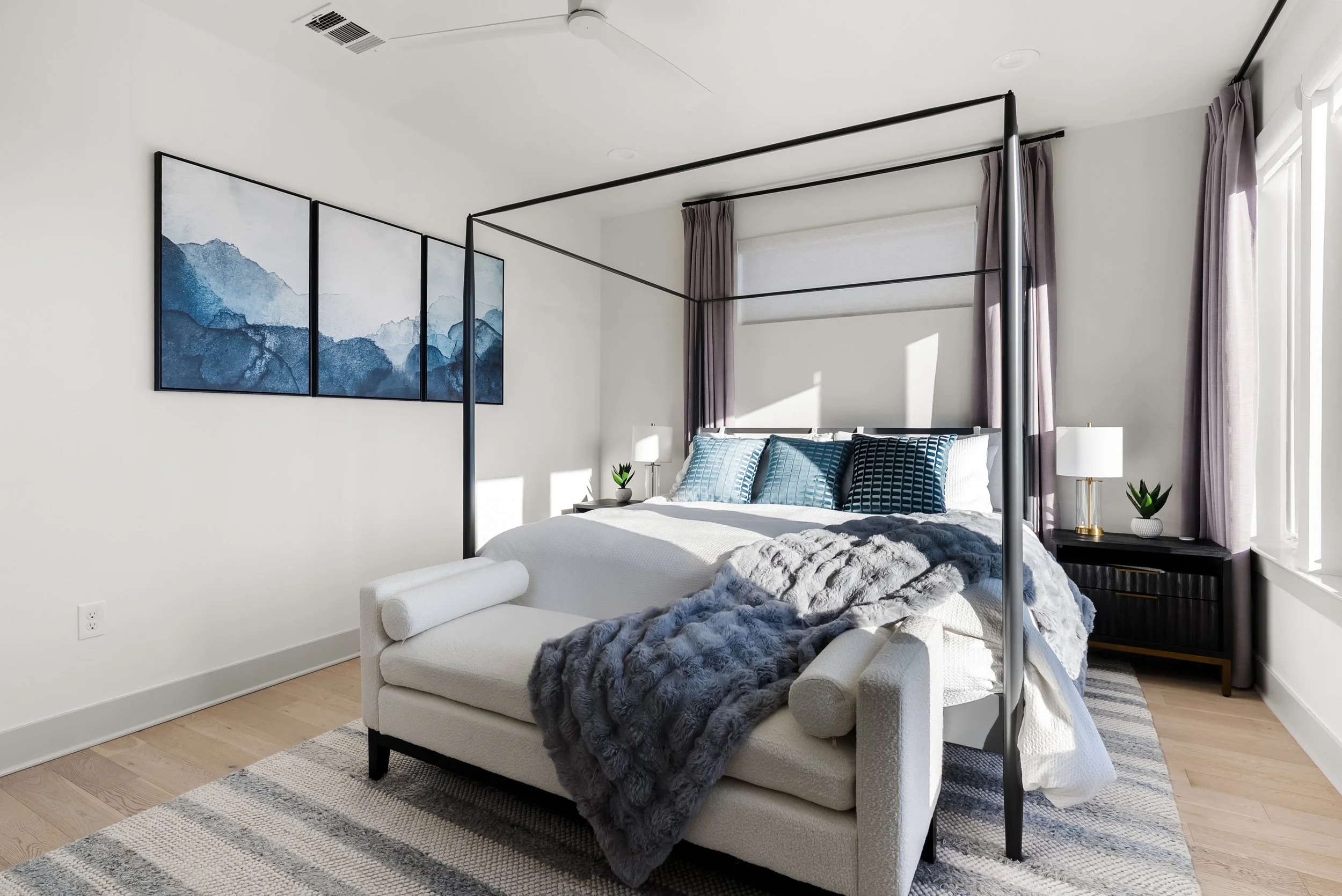 A bedroom with a white bed featuring blue and black pillows, a dark wood nightstand with a lamp and plant, lavender curtains, and a minimalist mountain art triptych on the wall.
