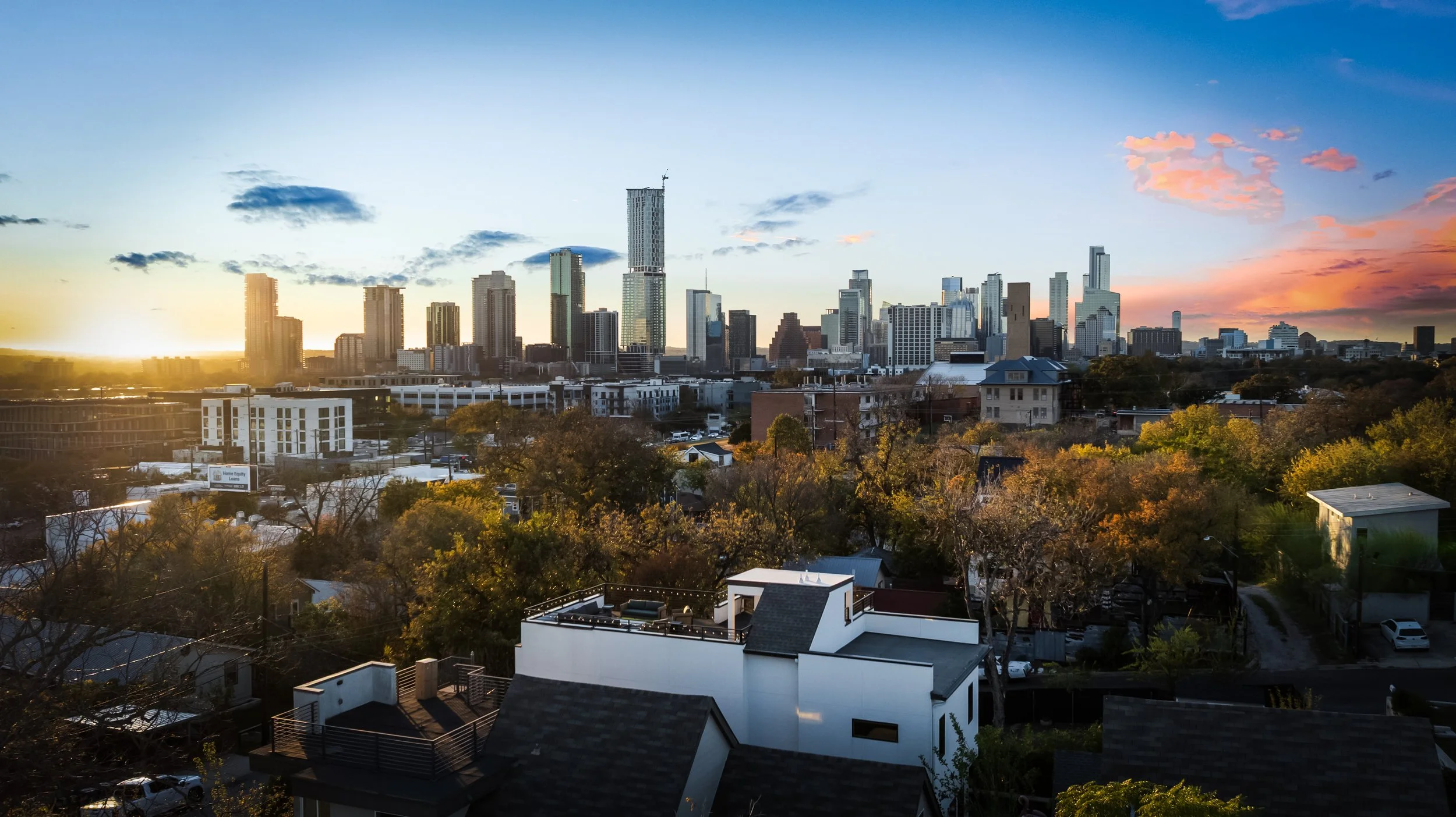 Sunset over a city skyline with tall buildings, trees in the foreground, and a dramatic colorful sky with pink clouds.