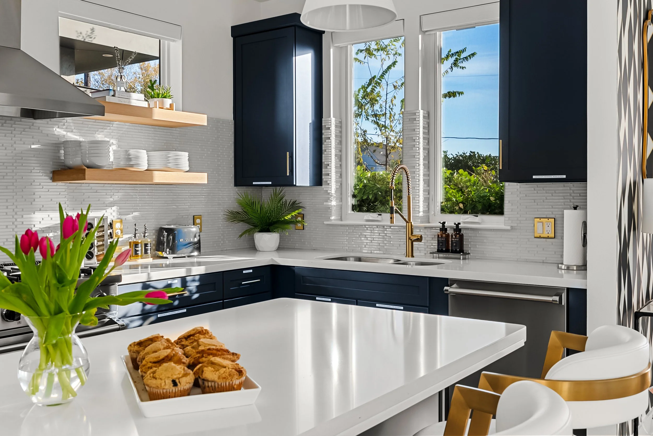 Modern kitchen with white countertops, navy blue cabinets, and a white island. A vase with pink tulips and a plate of muffins are on the island. Open shelves with white dishes, a toaster, and a plant are visible, along with a window showing greenery outside.