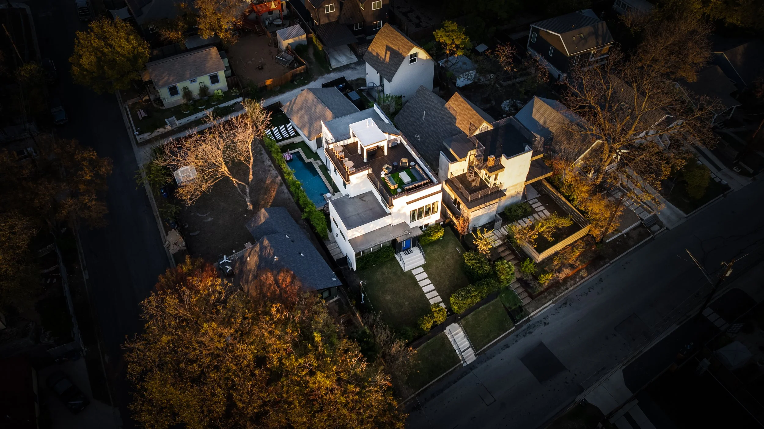 Aerial view of a modern house with a backyard pool, surrounded by other homes and trees, taken during sunset or early evening.