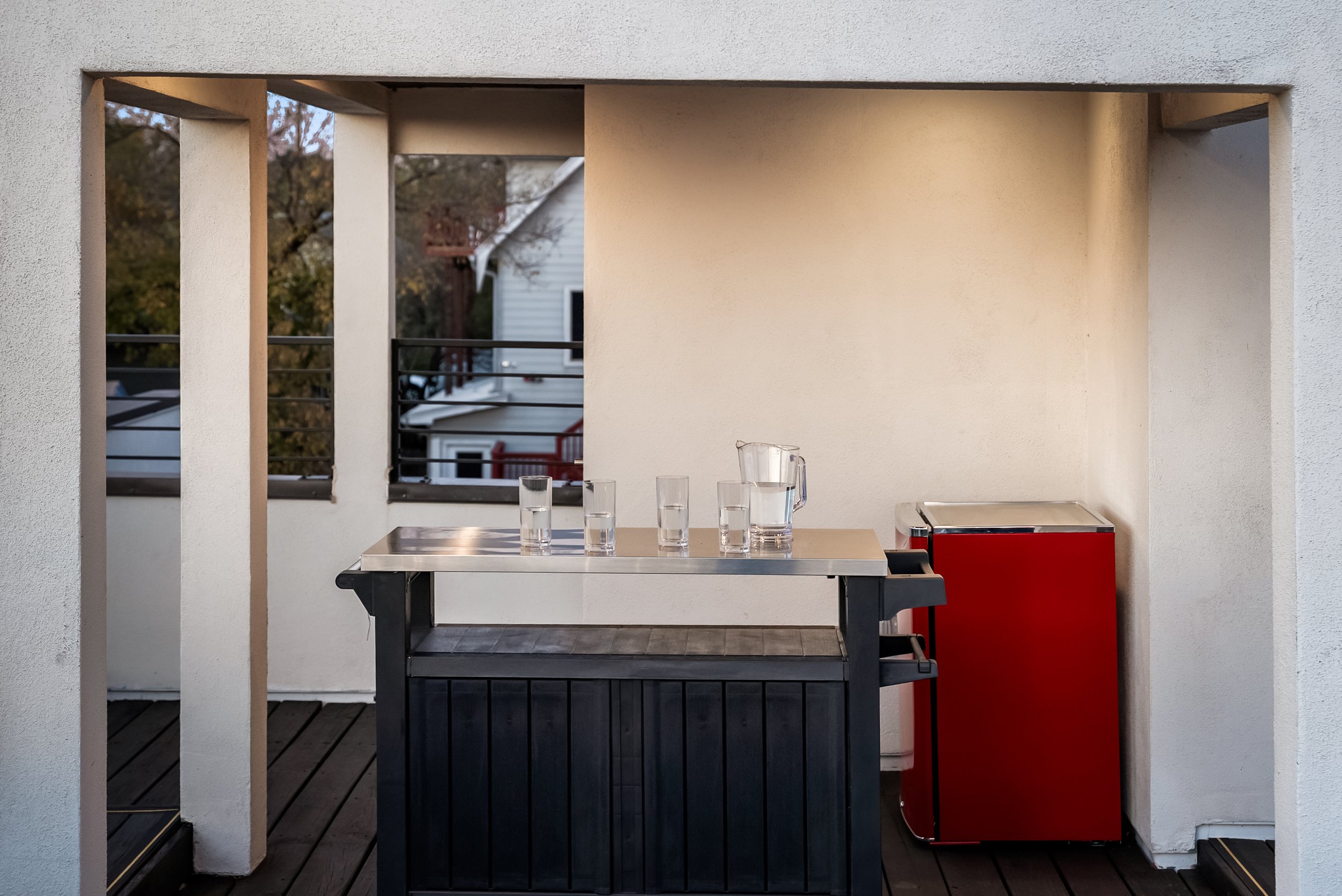 Outdoor patio with a black and stainless steel bar cart, four shot glasses, a glass pitcher, and a red mini fridge against a white wall with a balcony railing in the background.