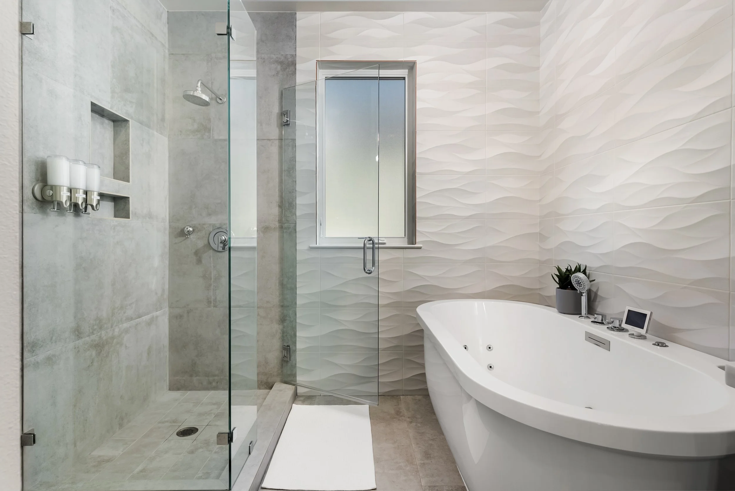 Modern bathroom with a glass-enclosed shower, a window, a white bathtub with a potted plant, a small digital clock, and decorative textured wall tiles.
