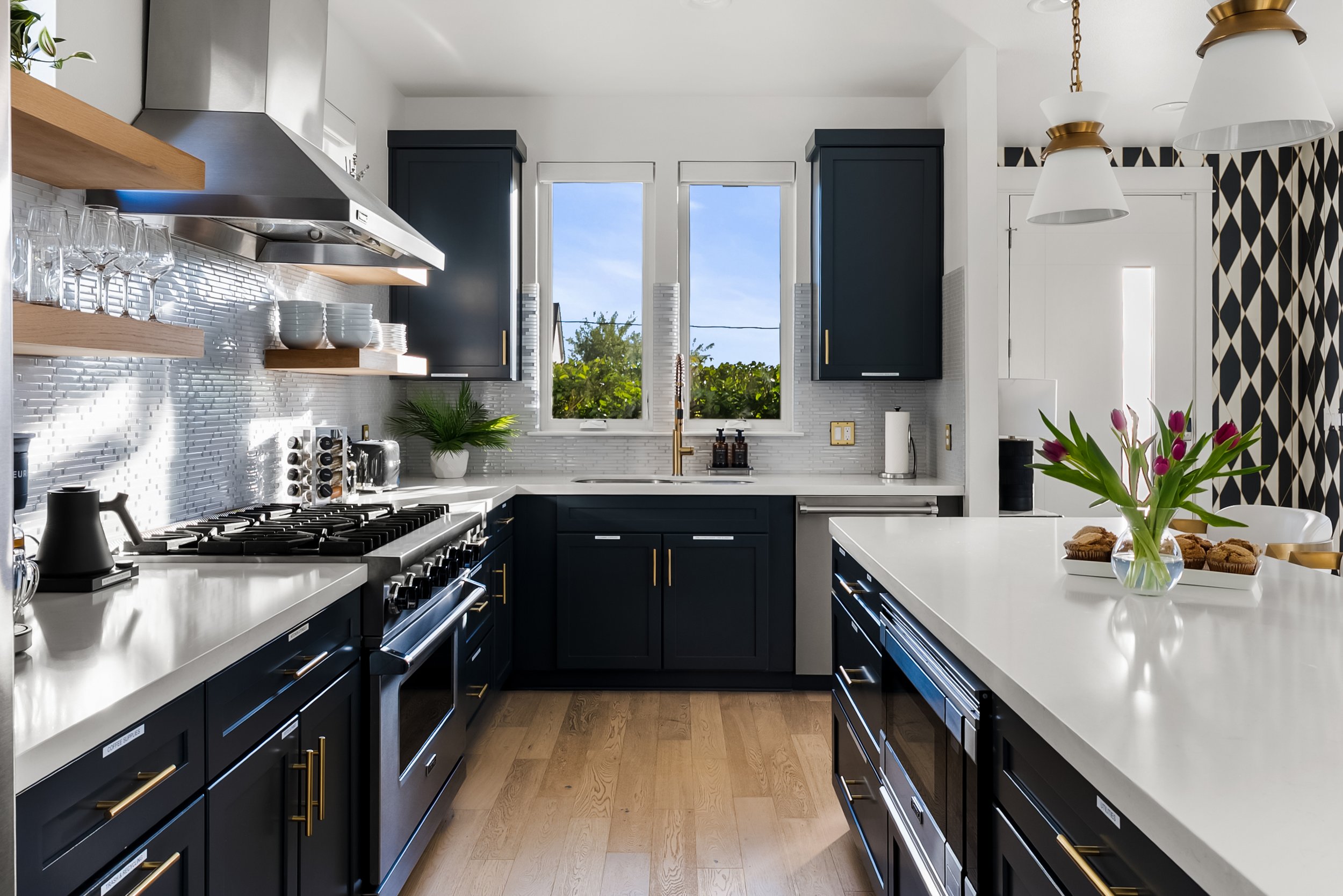 Modern kitchen with black cabinets, white countertops, and a white backsplash. Contains a stainless steel stove, shelves with glassware and bowls, a potted plant, and a window showing outdoor greenery. There are flowers on the island counter and modern lighting fixtures.
