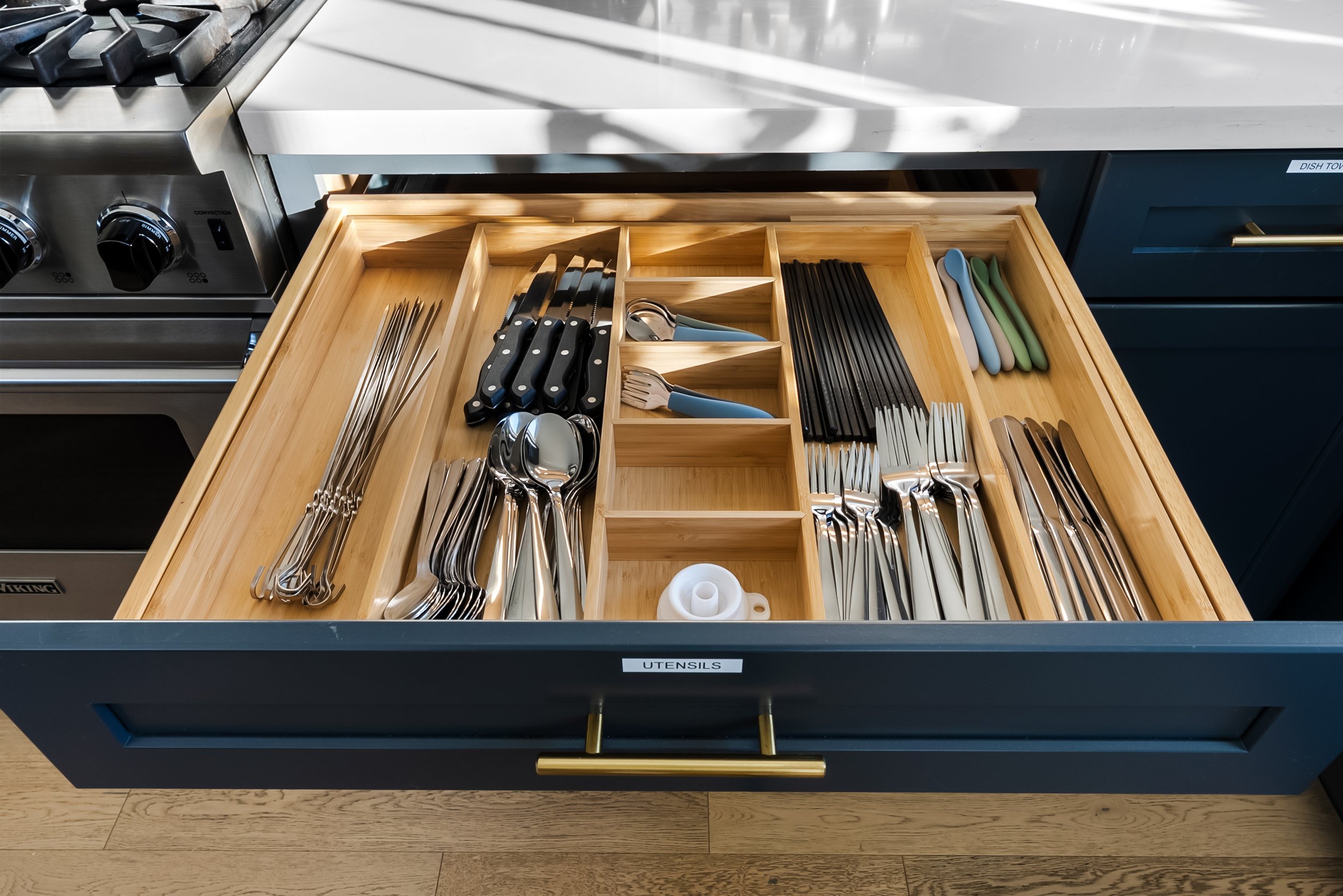 Open kitchen drawer organized with utensils, including knives, forks, spoons, and other kitchen tools, inside a wooden drawer with labeled compartments.