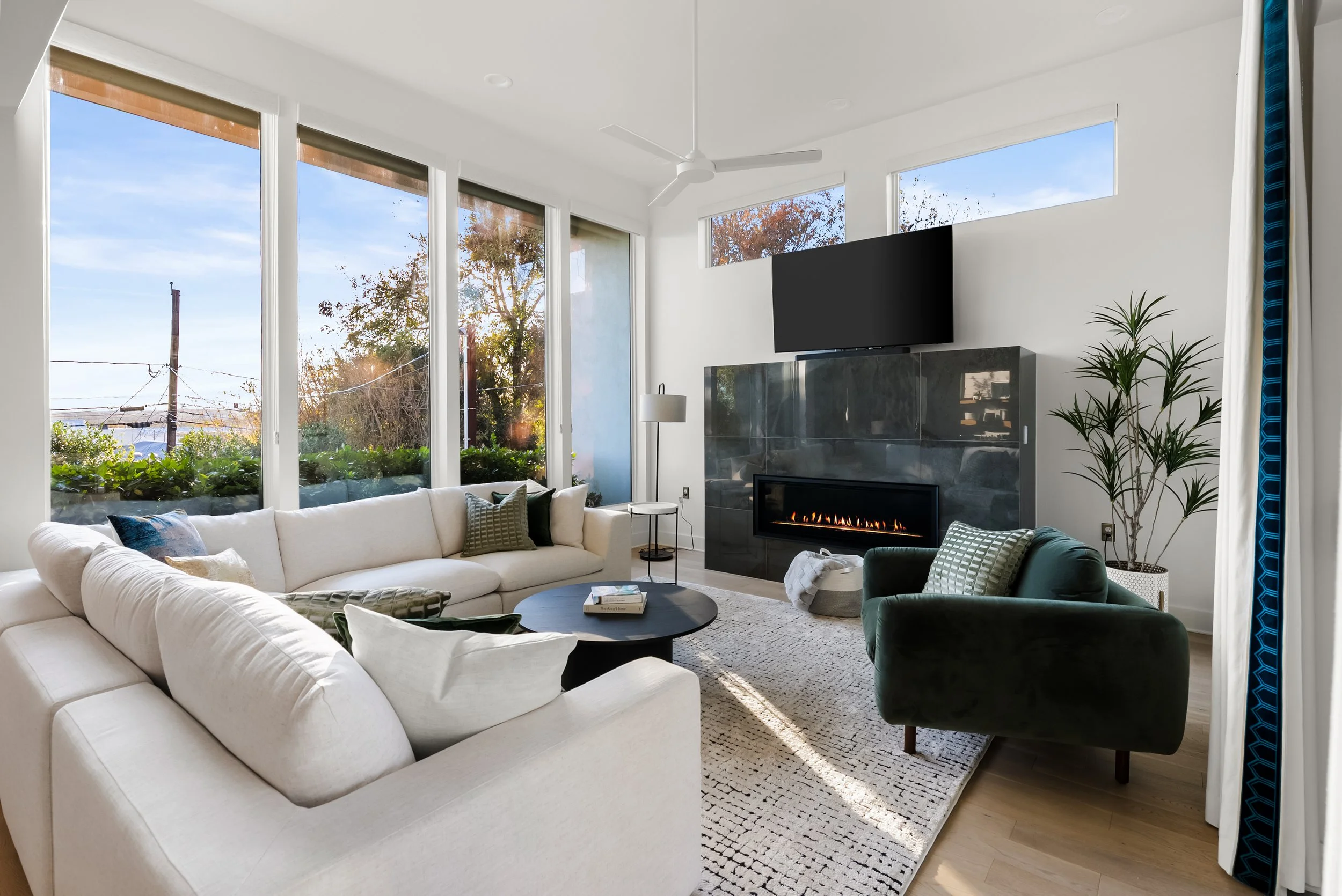 Living room with large windows, white couch, green armchair, black coffee table, fireplace, flat-screen TV, potted plant, and natural light.