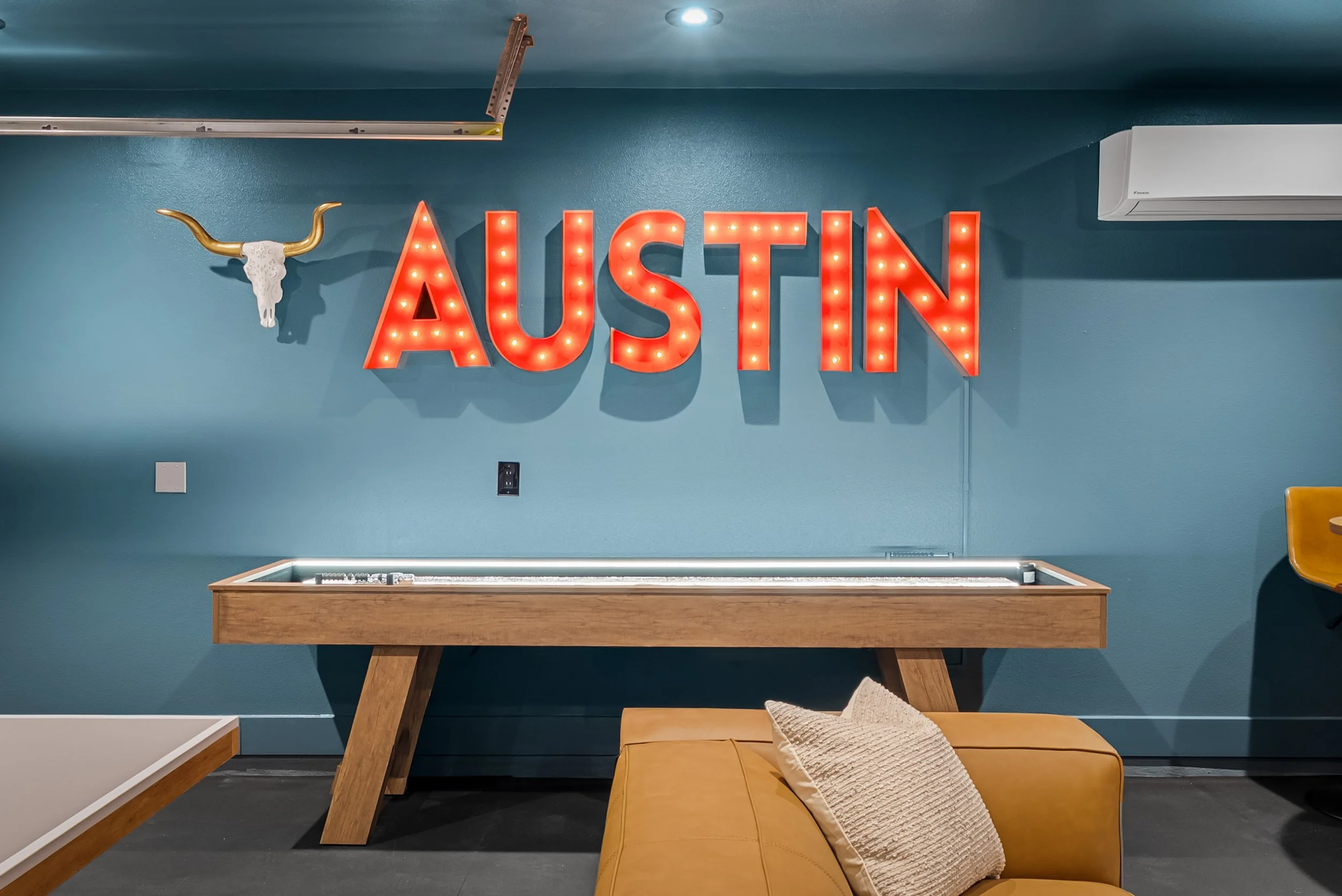 Decorative sign spelling 'AUSTIN' in red illuminated letters on a blue wall, with a mounted longhorn skull to the left and a blue air conditioning unit on the right.