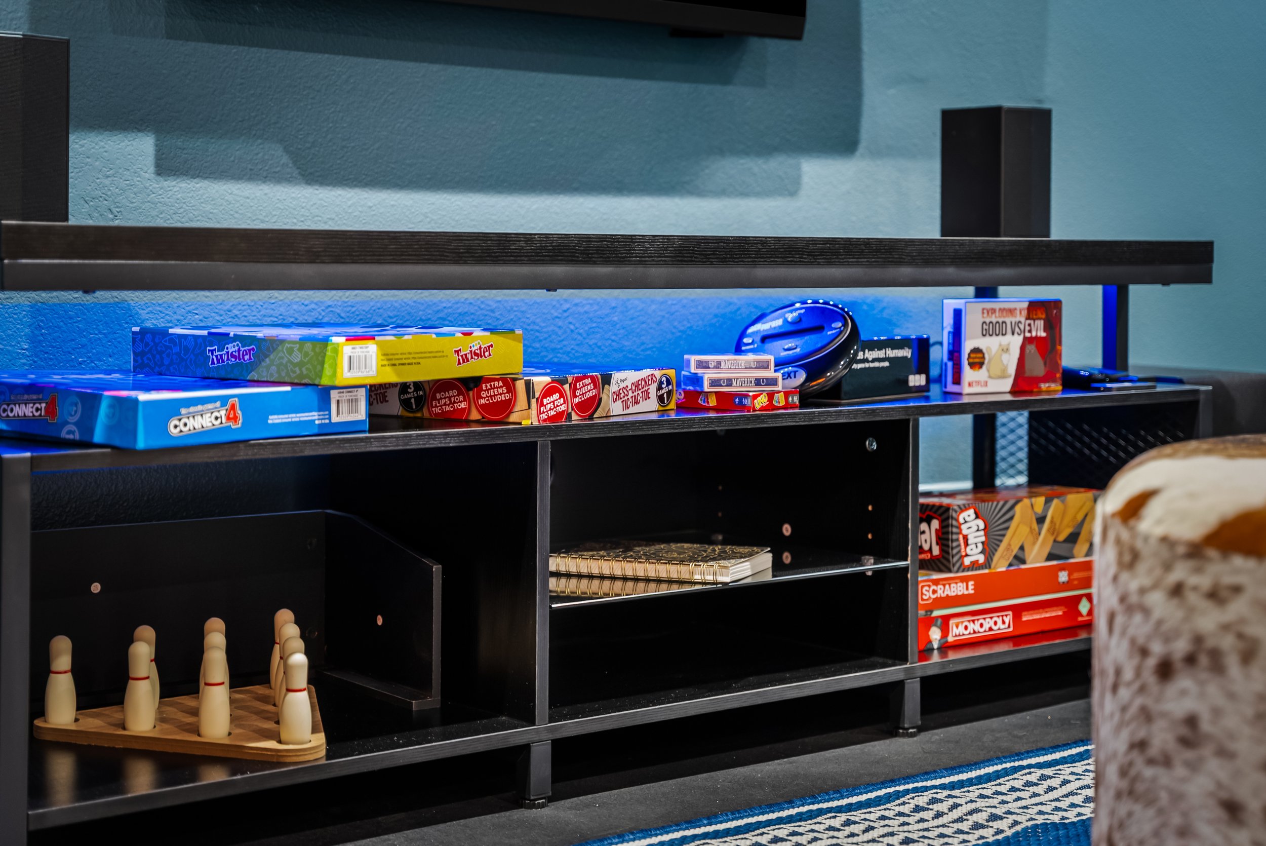 Game shelf with board games, a game console controller, and a few movie cases, with a partial view of a bowling pin set in the foreground.