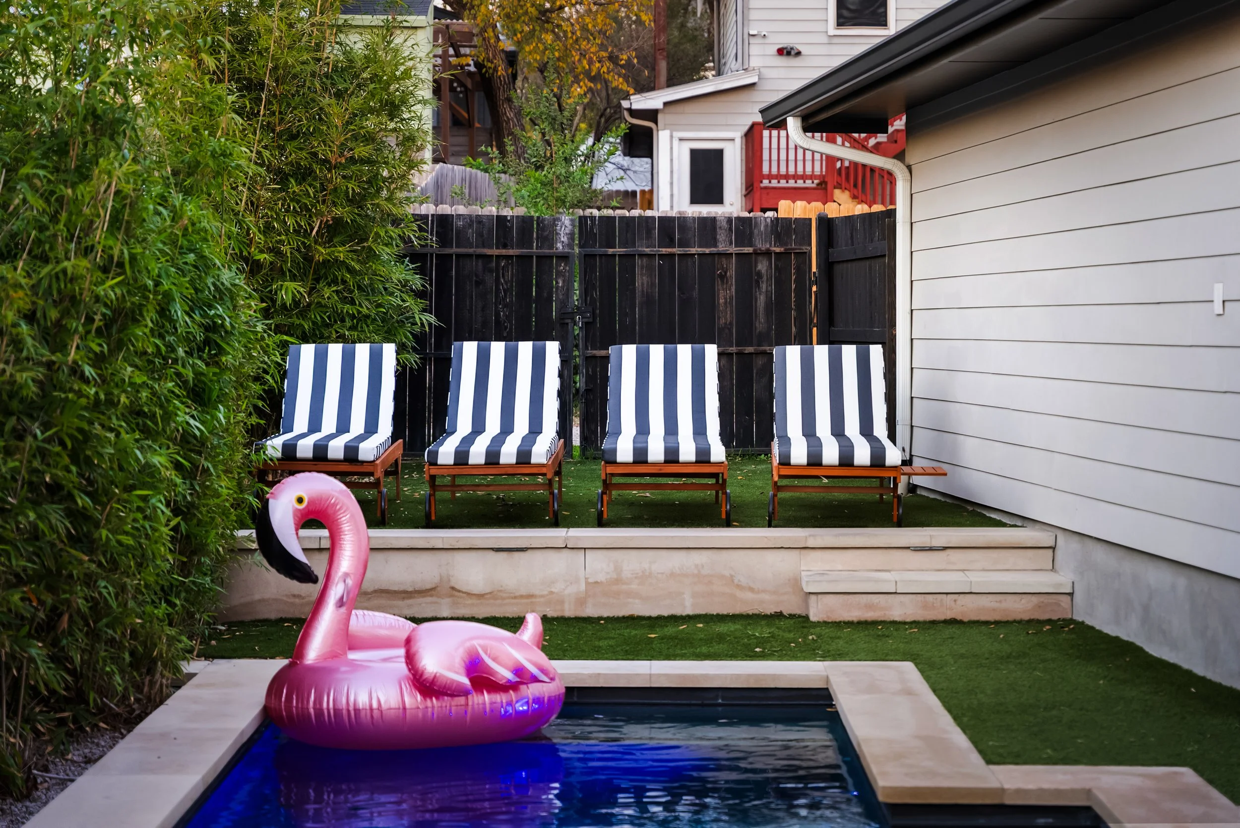Backyard with four striped lounge chairs, inflatable pink flamingo pool float, small swimming pool, and greenery on the left side, with a white house wall on the right and a black fence in the background.