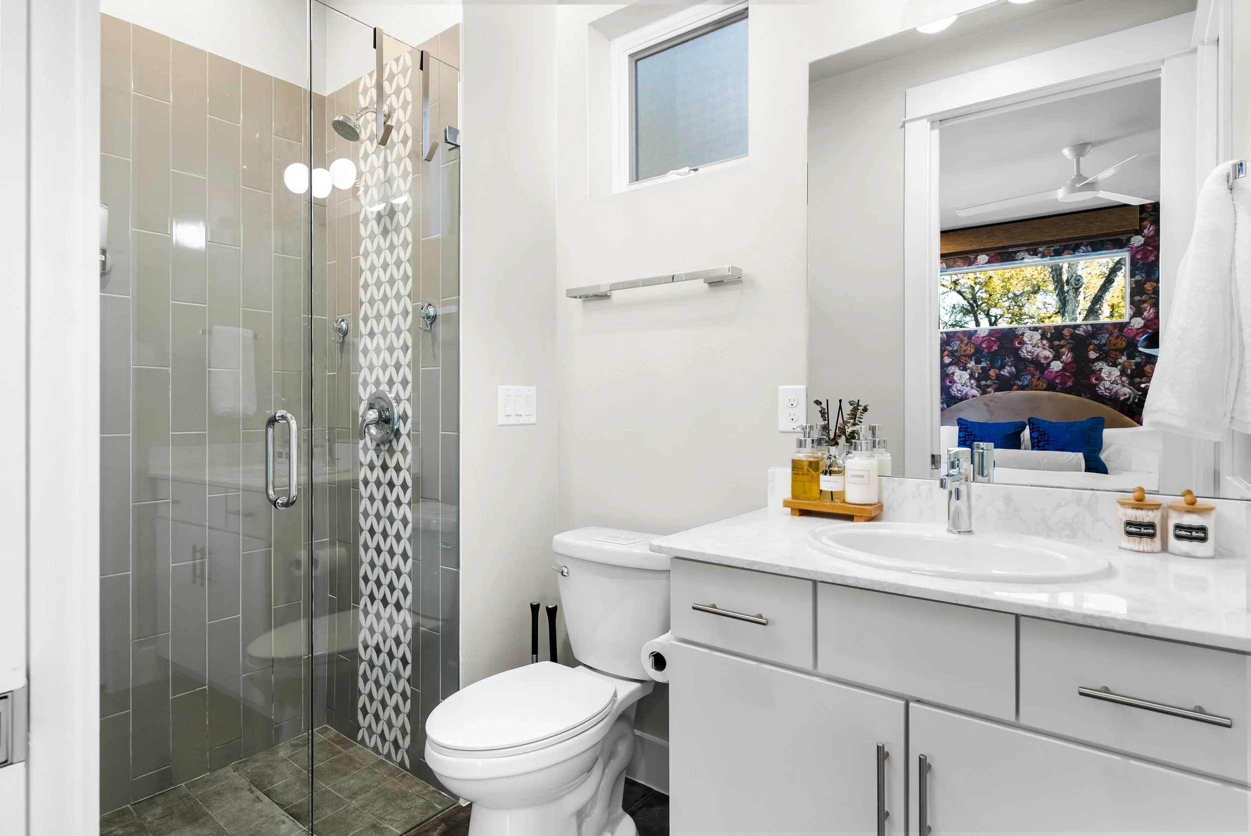 Modern bathroom with a glass shower, white toilet, white vanity with marble countertop, and a mirror reflecting a bedroom with floral wallpaper and a window showing trees outside.