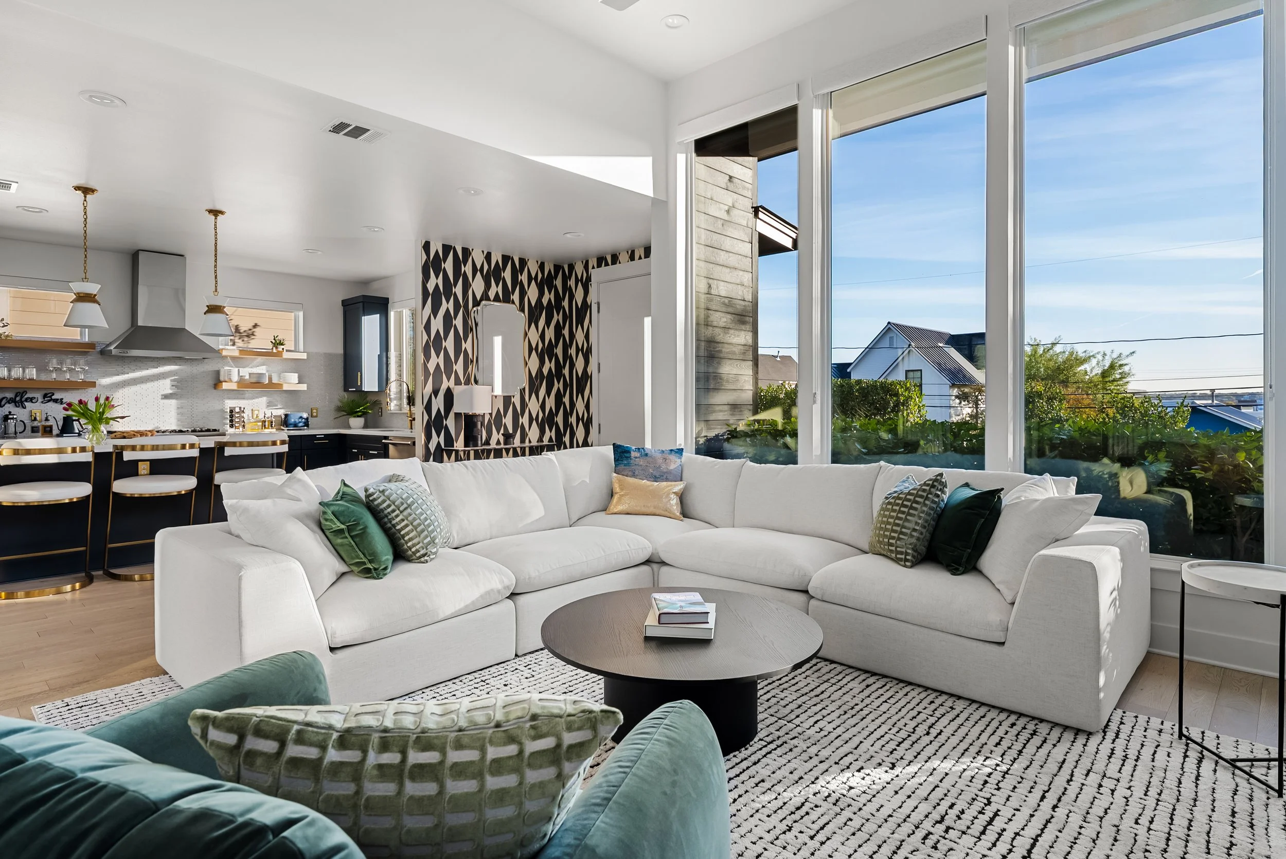 Living room with a white sectional sofa, various pillows, round coffee table, large windows showing a view of neighborhood houses, and a kitchen area with pendant lights and bar stools in the background.