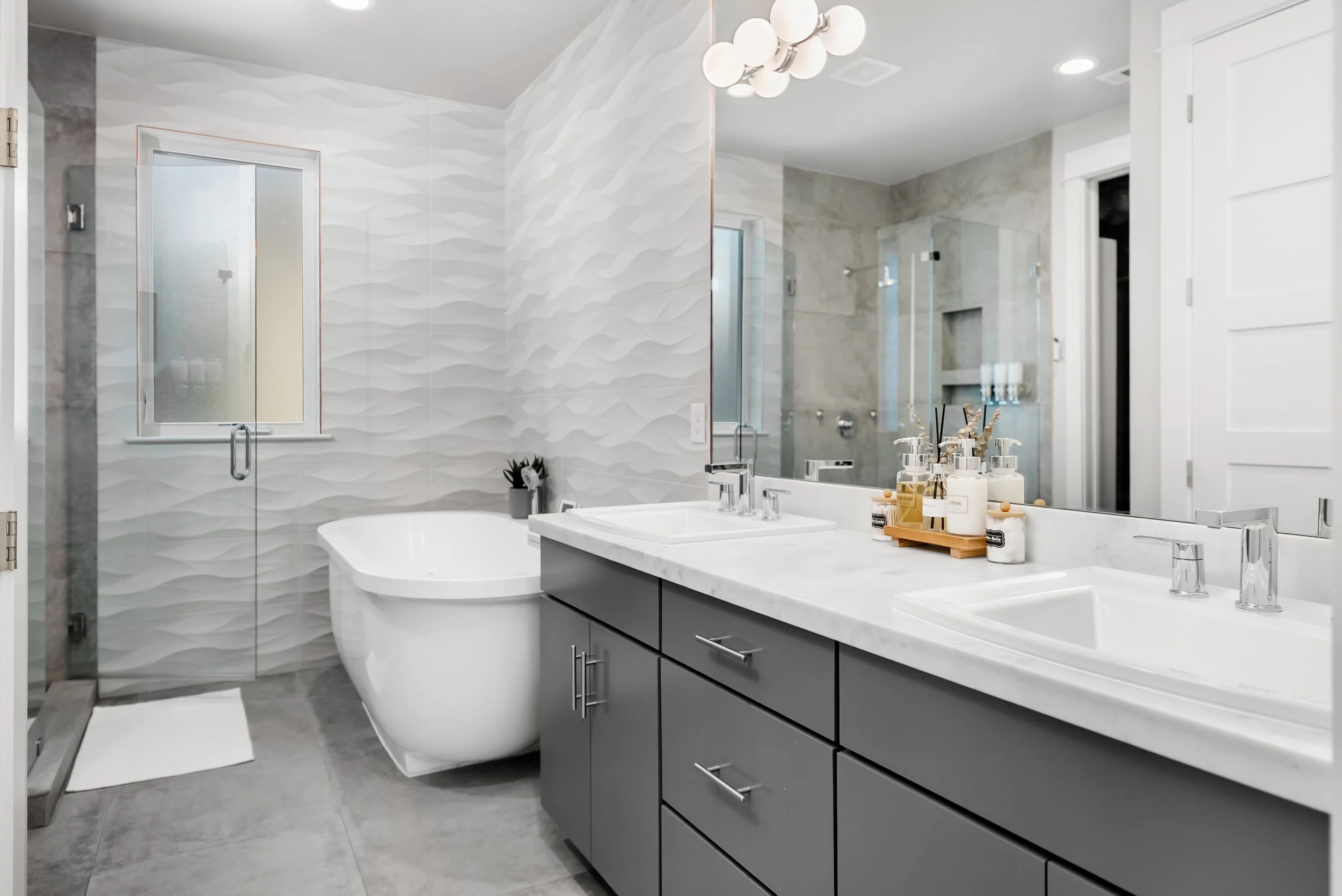 Modern bathroom with dual sinks, large mirror, grey cabinets, white countertop, decorative items, freestanding bathtub, window, glass shower with built-in shelves, textured grey wall tiles, and ceiling light fixtures.