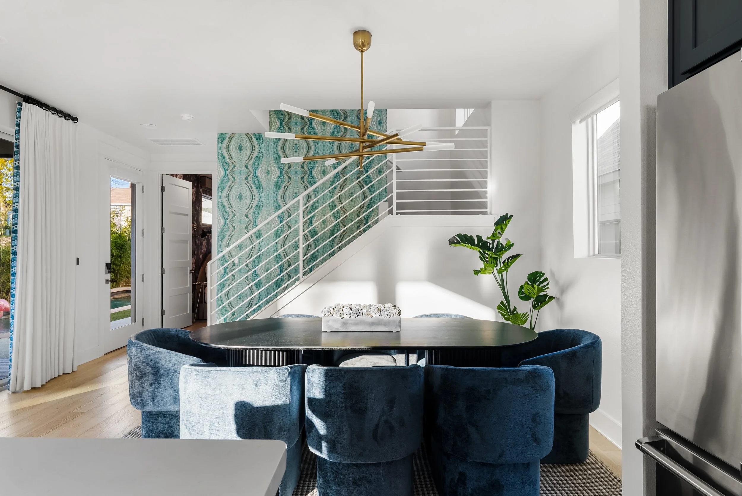 Modern dining room with a black oval table surrounded by blue velvet chairs, a gold and white chandelier, a green plant, and a staircase with a teal patterned wall.
