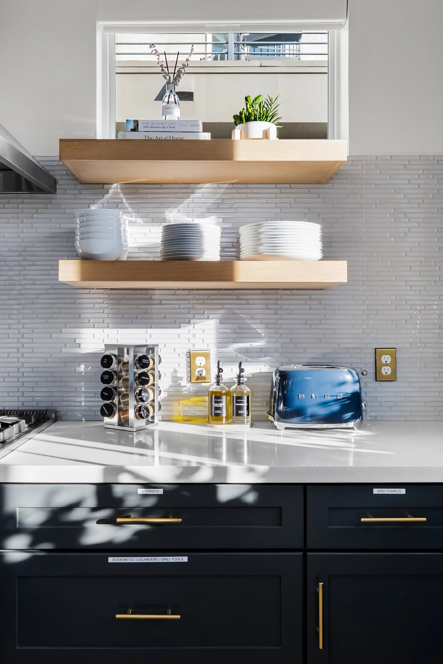 Kitchen with white countertop, black drawers with gold handles, open shelving with white dishes, a small window with sunlight and shadows, and various kitchen items like a spice rack, soap dispensers, a toaster, and some books.