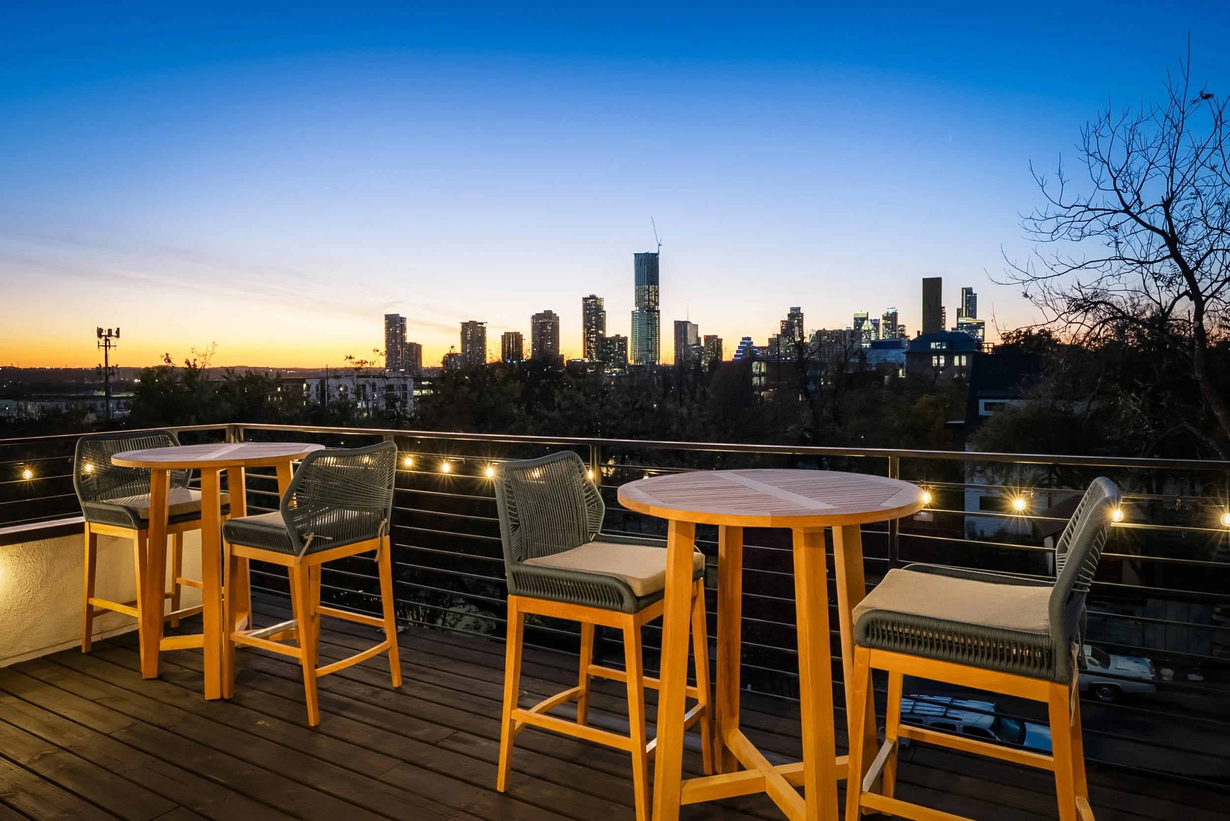 Outdoor balcony with wooden tables and chairs overlooking a city skyline at dusk, with string lights along the railing.