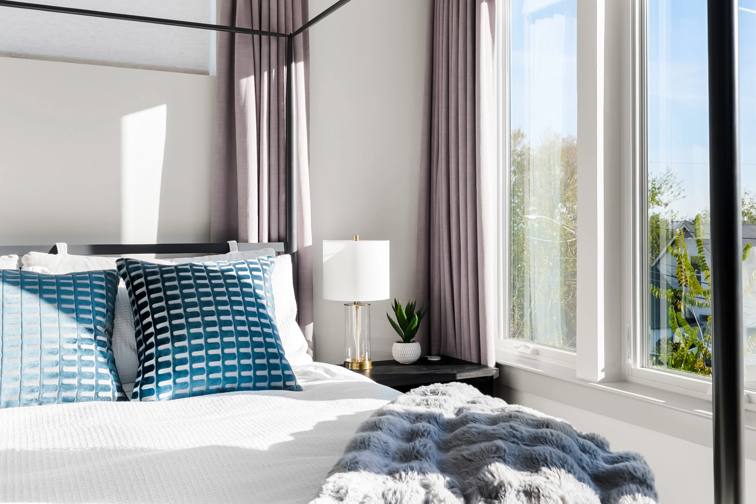 A bright bedroom featuring a bed with white bedding and blue patterned pillows, a nightstand with a white lamp, a potted plant, and a window with purple curtains letting in natural light.