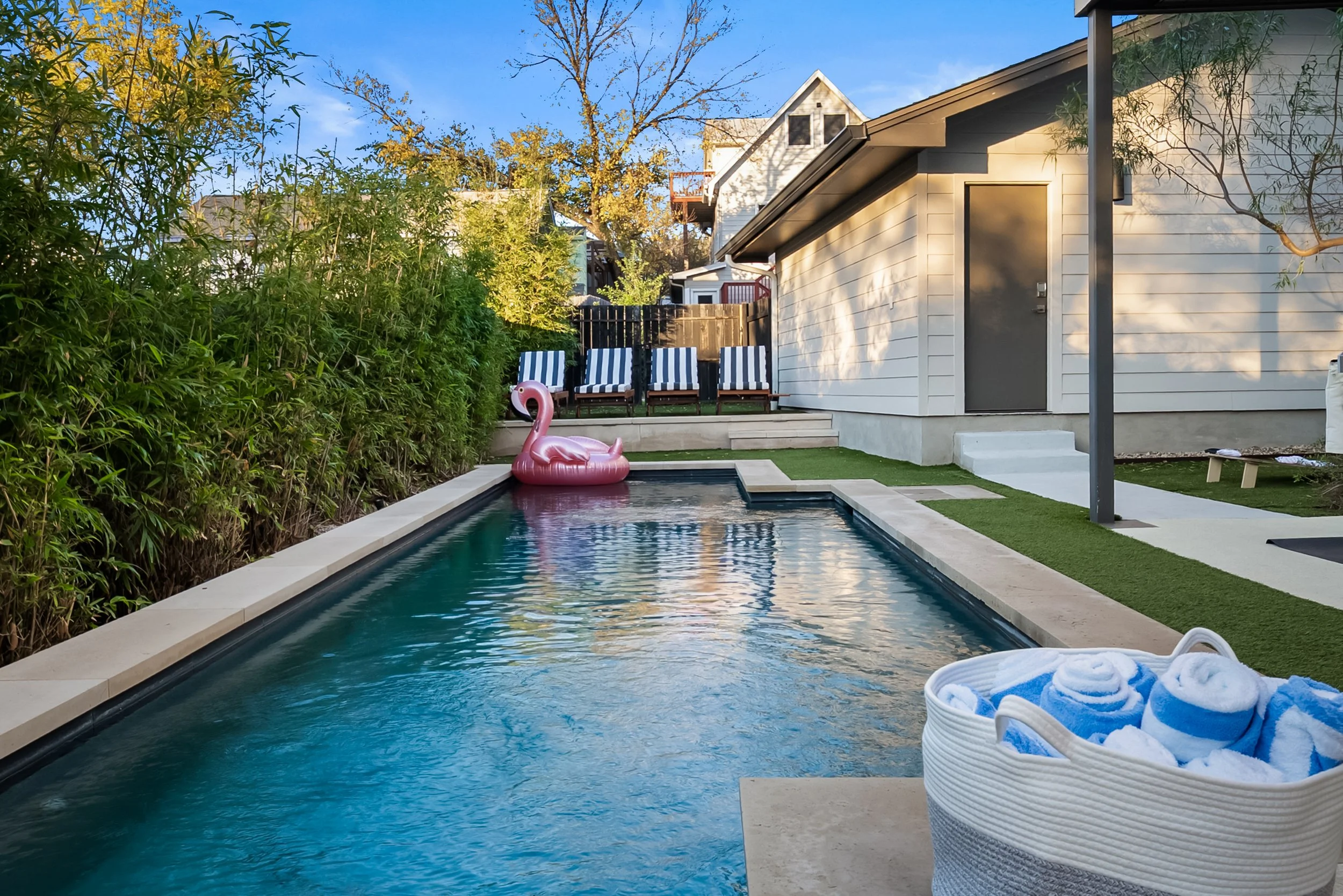 Backyard with a rectangular swimming pool, inflatable pink flamingo float, basket of rolled white towels, and outdoor seating with striped cushions on the patio. There are lush green bushes on the left and a house with white siding on the right.