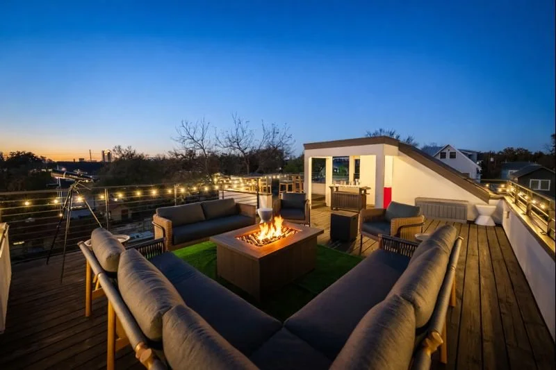 A rooftop patio during dusk with outdoor seating, string lights, a telescope, and cityscape view.