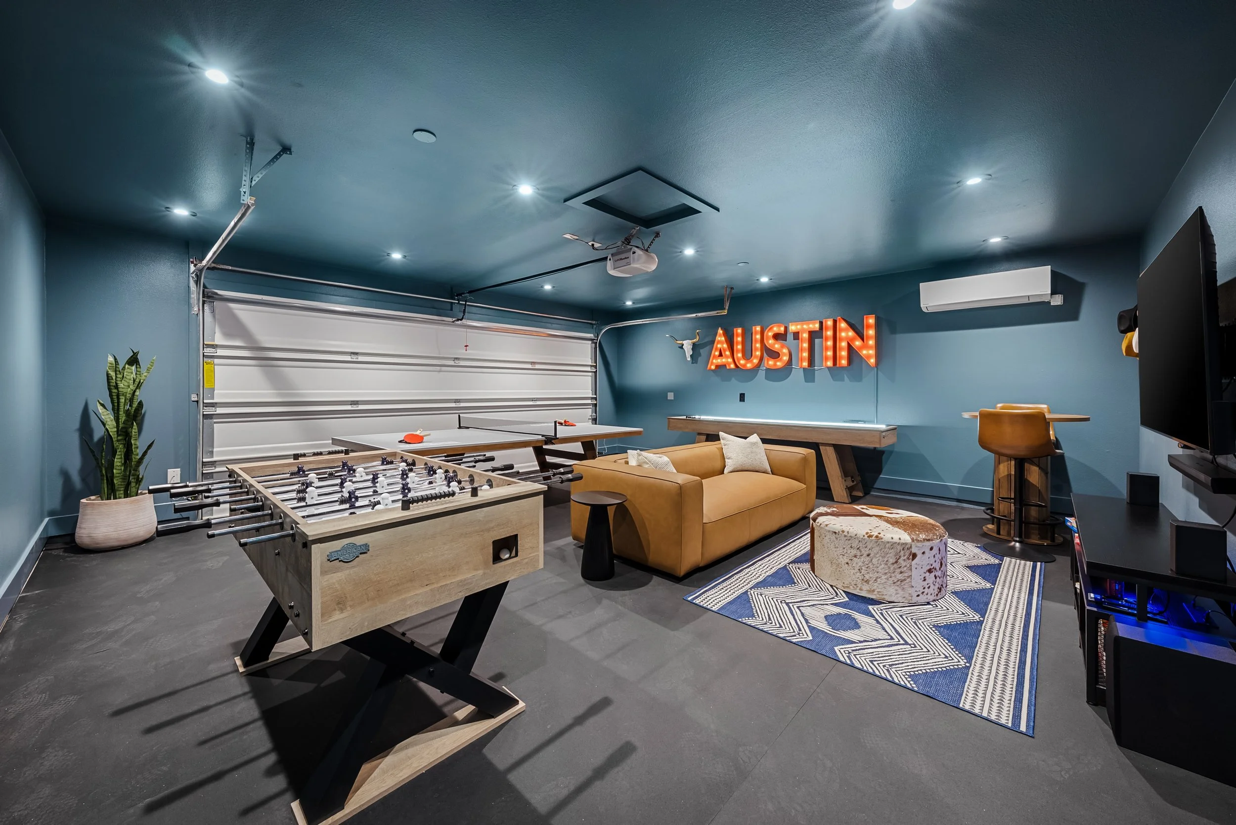 A modern basement game room featuring a foosball table, ping pong table, a beige sofa, a cowhide ottoman, a blue and white rug, a wall sign spelling "AUSTIN" with lights, a television, a tall plant, an air conditioning unit, and a barstool.