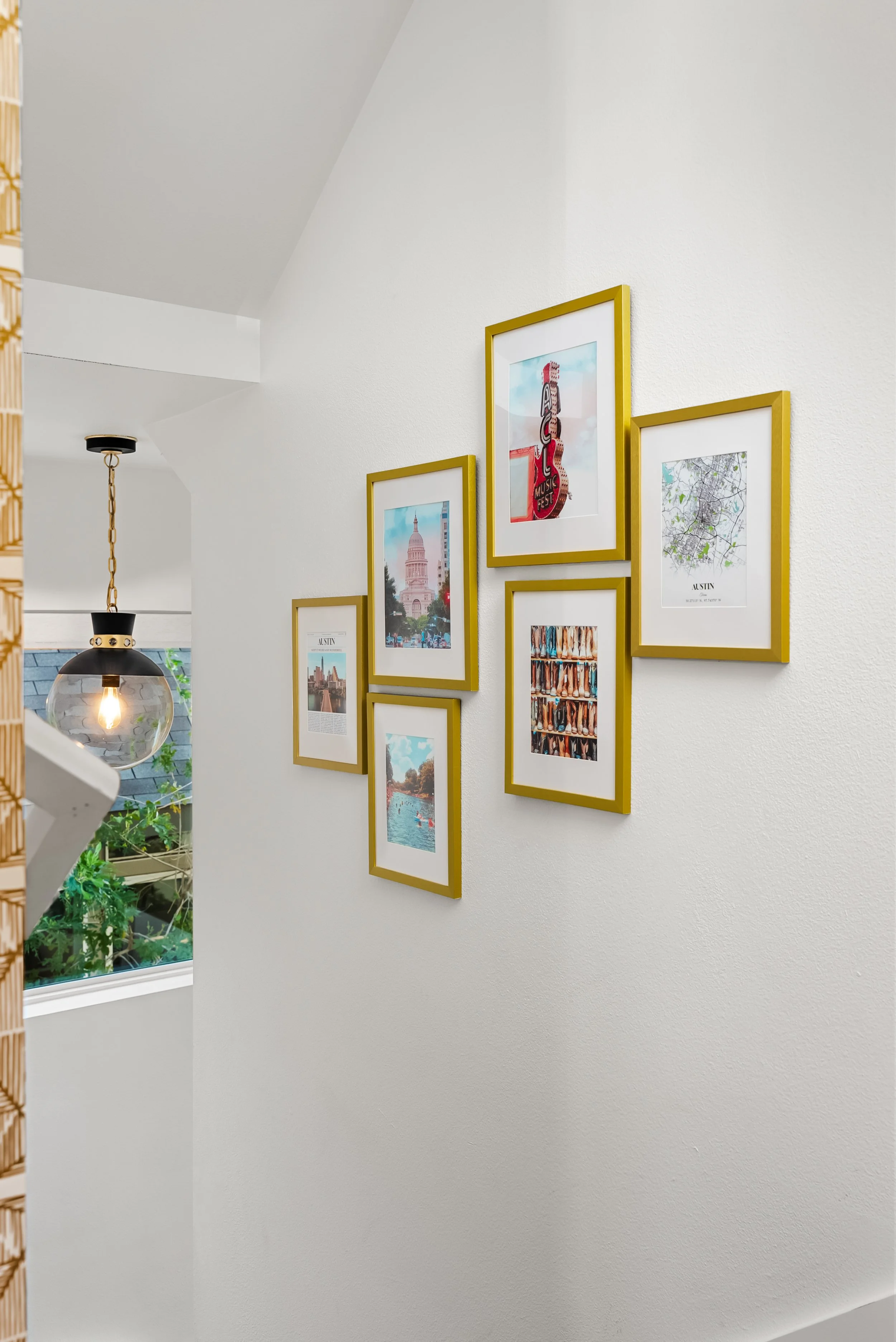 Interior wall decorated with a gallery of seven framed pictures with gold borders, including landmarks, cityscapes, and artwork, with a hanging light fixture and a window with greenery