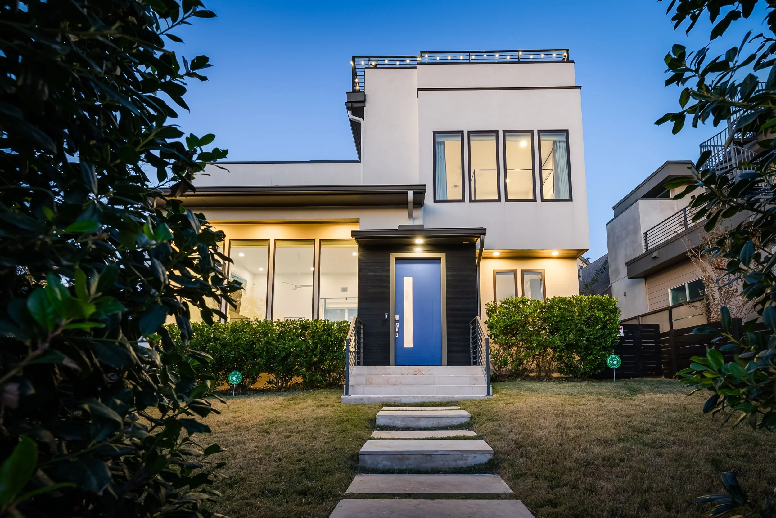 Modern two-story house with a blue front door, illuminated by exterior lights, surrounded by a neatly maintained lawn and bushes, during twilight.