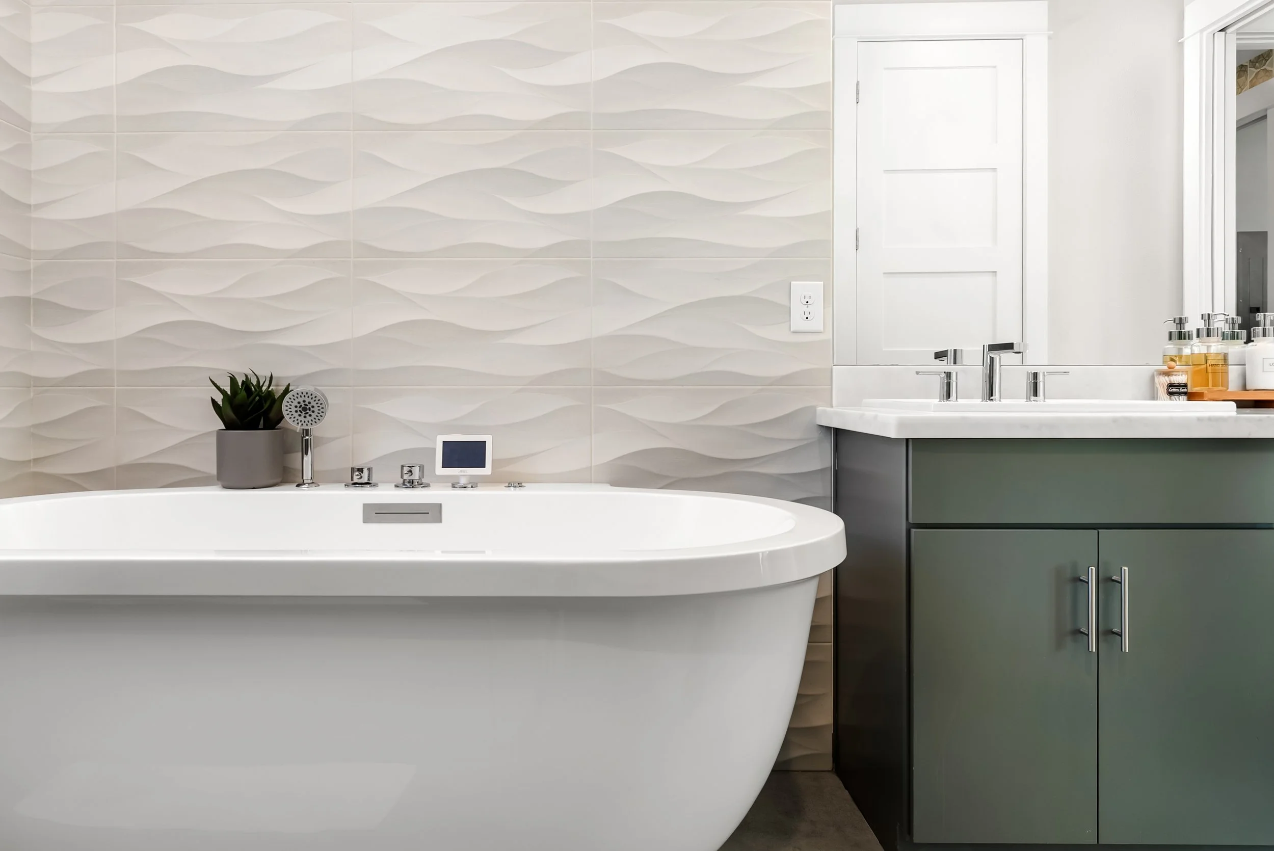 Modern bathroom with a white bathtub, a green vanity with a white countertop, a faucet, and an assortment of soap dispensers. A plant and a digital control panel are placed on the bathtub edge, with textured wall tiles and a mirror in the background.