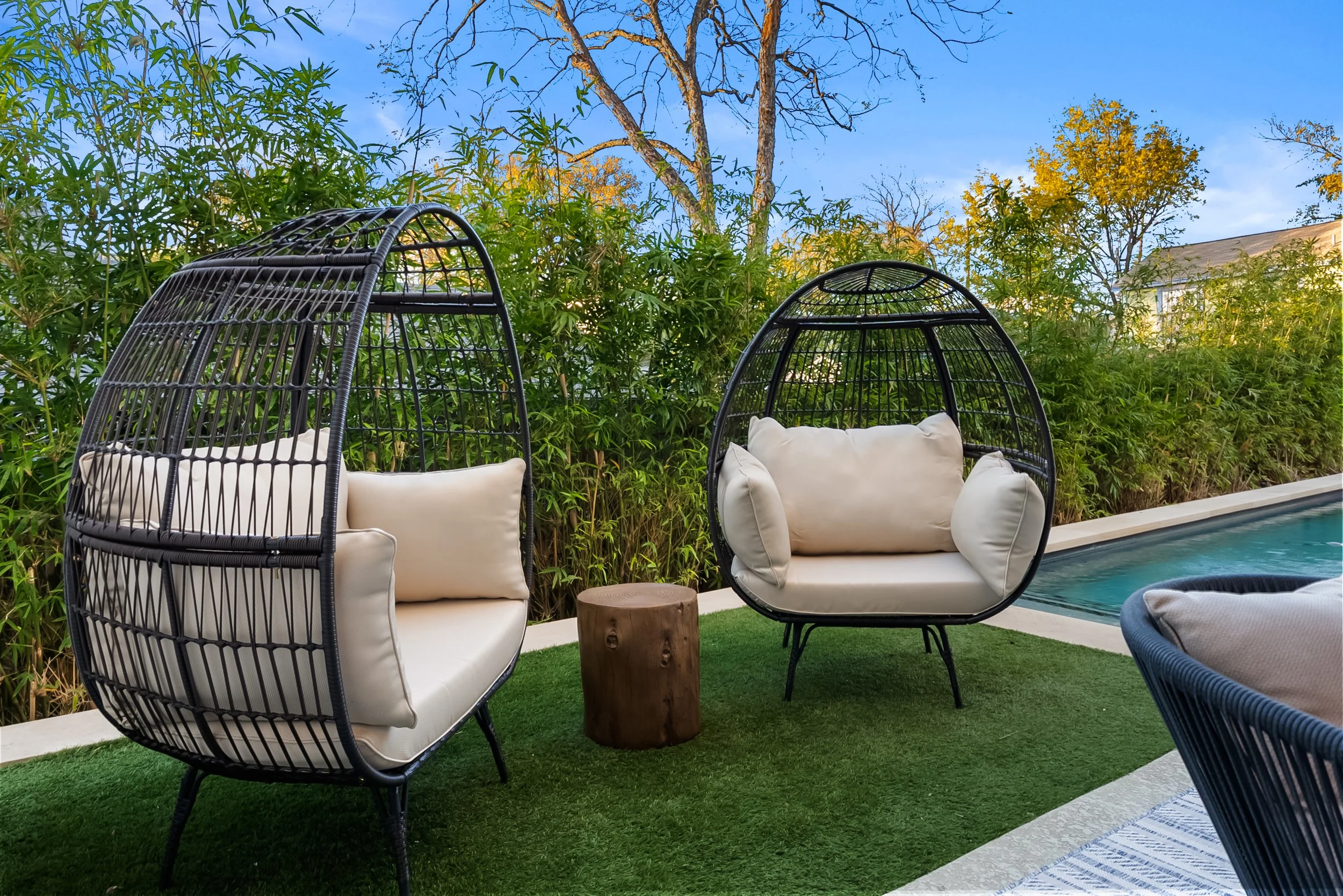Outdoor patio scene with two modern black rattan hanging chairs with white cushions, a small wooden stump table, and part of a swimming pool. Green bushes and trees in the background under a clear blue sky.
