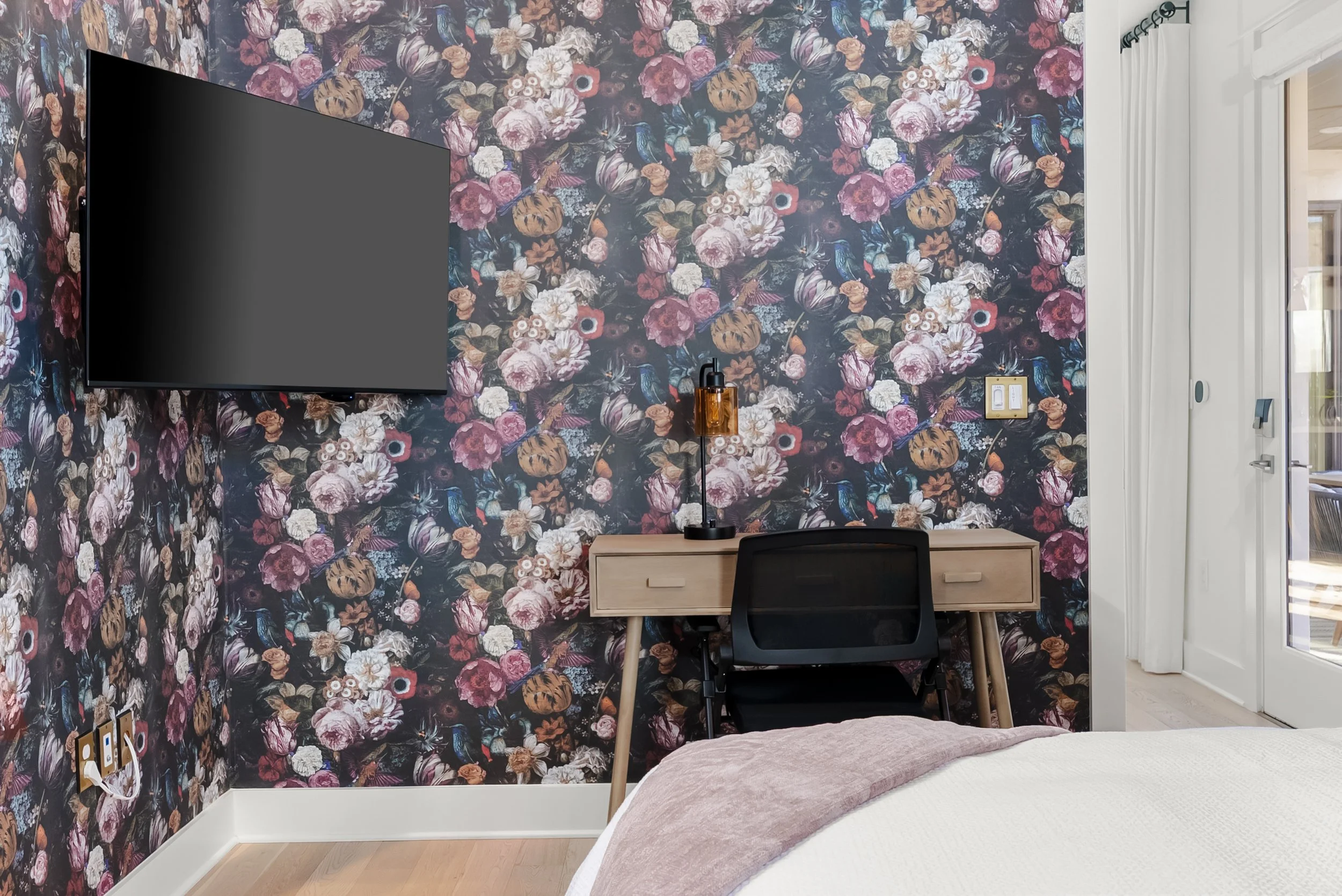 A floral accent wall in a bedroom with a mounted flat-screen TV, a wooden desk with a black chair, a decorative table lamp, and white curtains over a glass door.