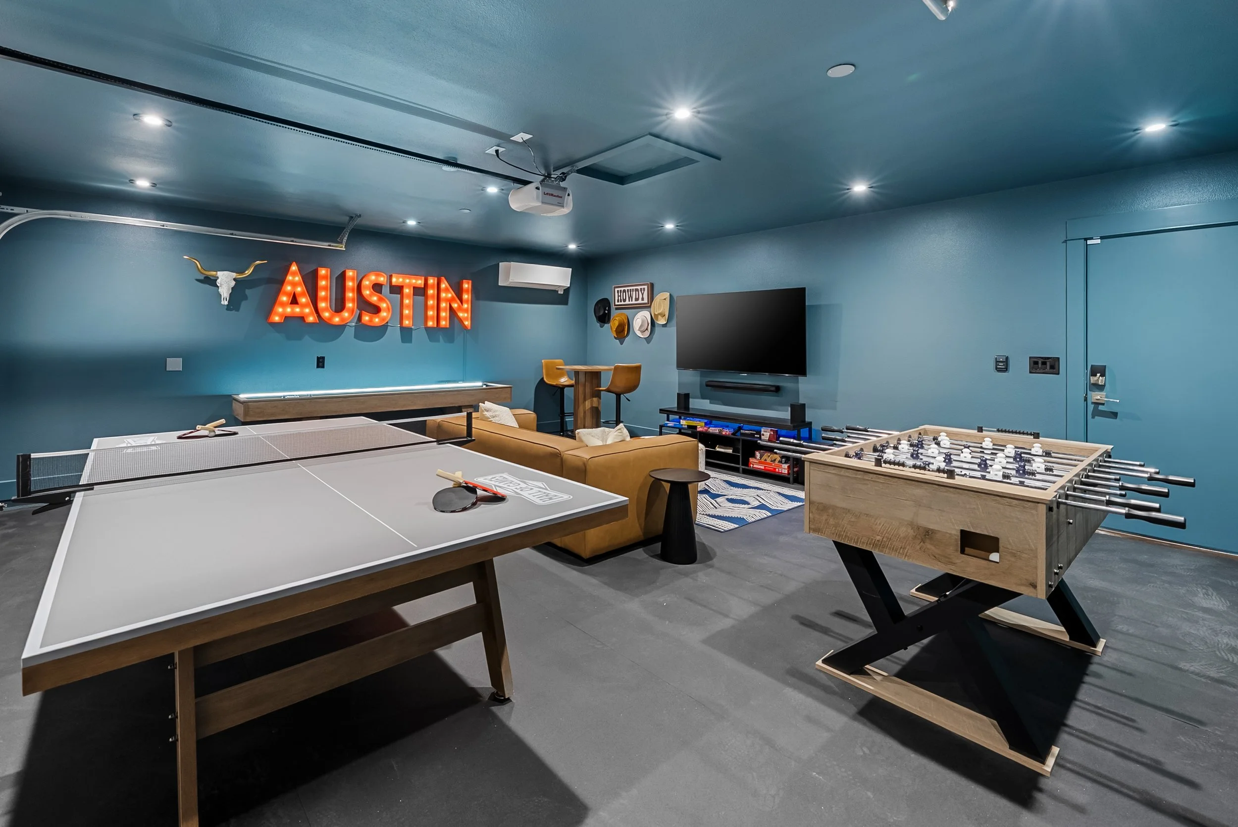 A game room with a ping pong table, foosball table, a sofa, a large TV on the wall, and decorative signs including a red neon sign that says 'AUSTIN' with a bull skull and horns above it.
