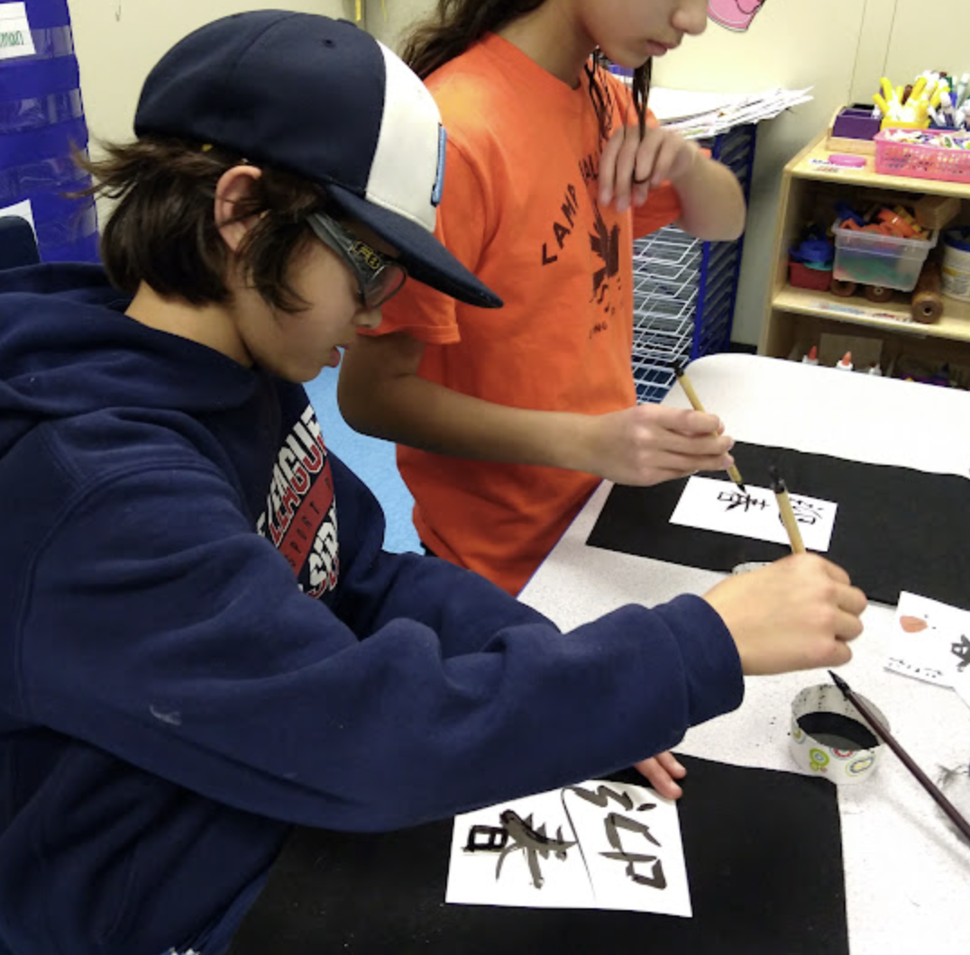 Two children working on calligraphy at a table, with art supplies and paper in a classroom setting.