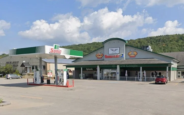 Station-service avec enseignes Sonic, Coop, et Délifrance, face à un centre commercial ou supermarché dans une zone rurale avec des collines en arrière-plan.